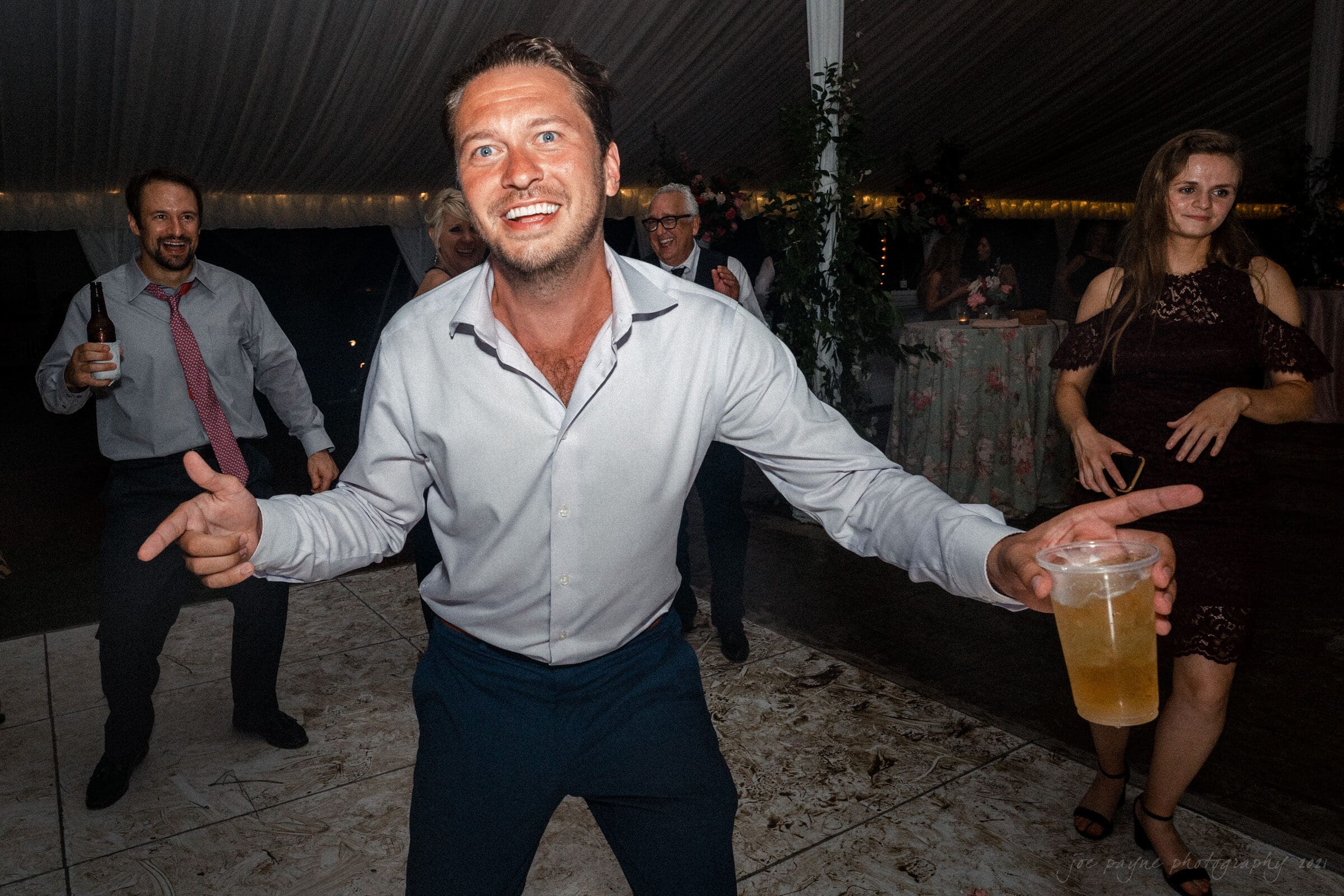 A man in a light shirt and dark pants dances energetically at a party, holding a drink in one hand. Other guests, casually dressed, dance and socialize in the background under a decorated tent.