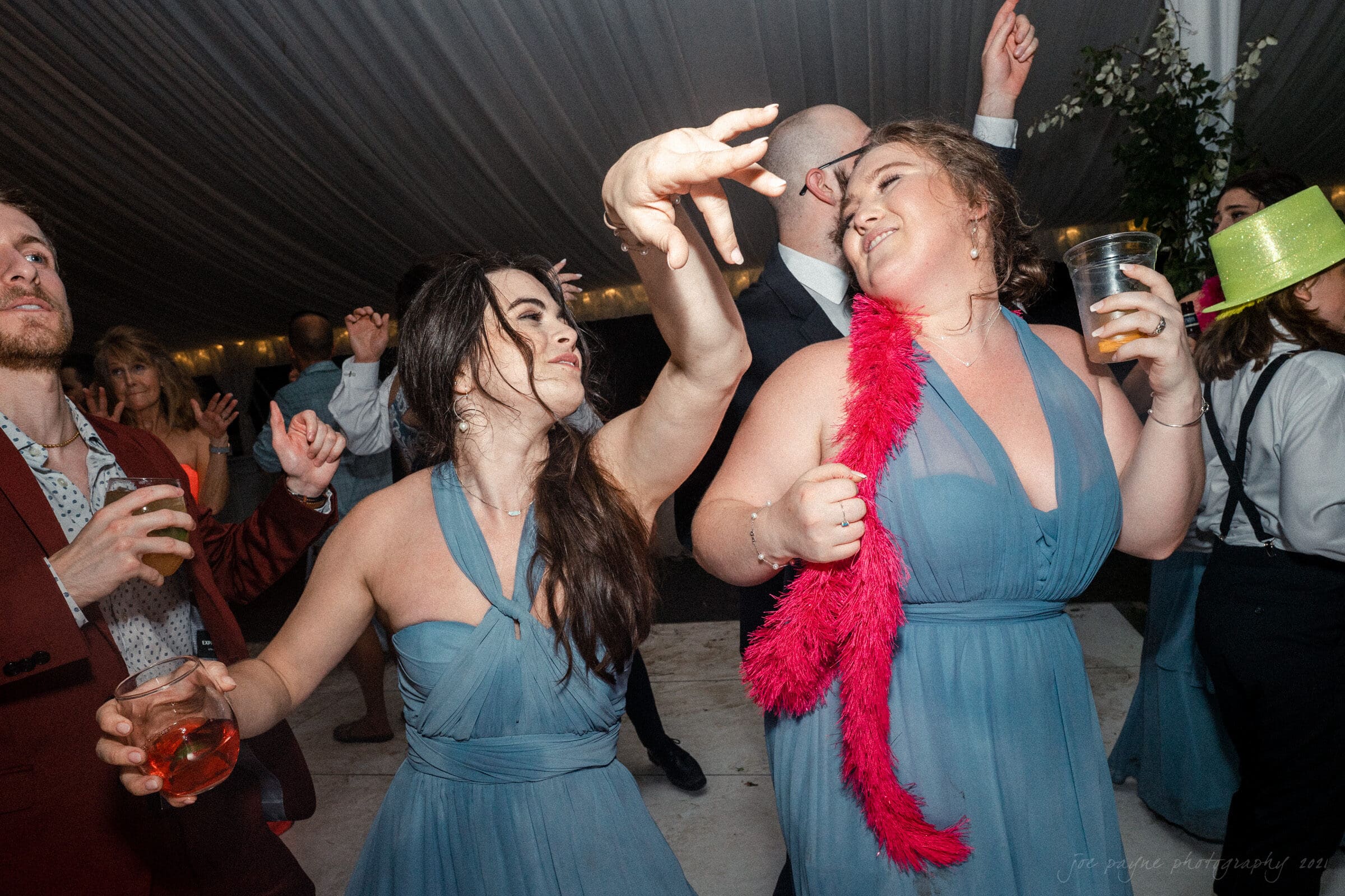 A lively dance scene at a party with people in formal attire. Two women in blue dresses energetically dance, with one wearing a pink feather boa. Others around them are also dancing under a white tent ceiling.