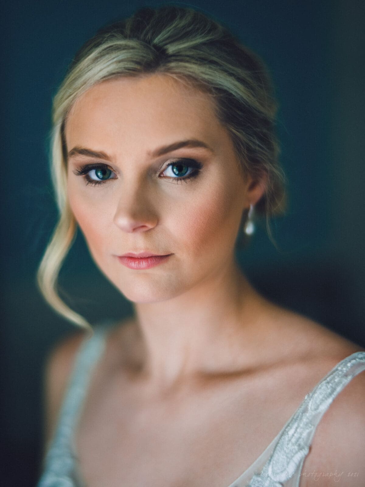 A woman with blonde hair and wearing a white bridal dress looks directly at the camera. Her hair is styled up with a loose strand framing her face, and she wears pearl earrings. The background is softly blurred.