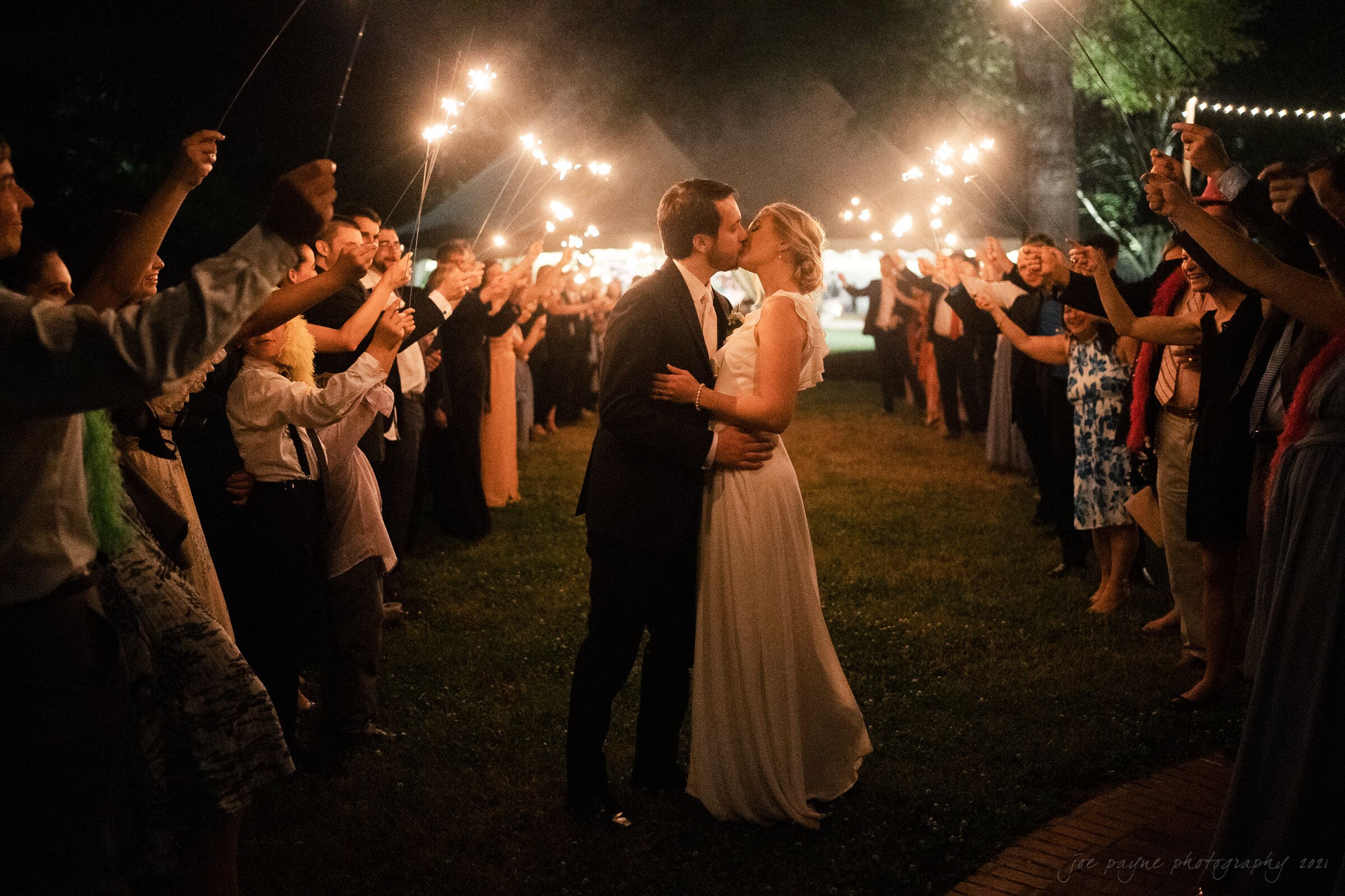 A bride and groom share a kiss on a grassy path, surrounded by guests holding sparklers. The scene is illuminated by string lights and the night sky, creating a warm and festive atmosphere.