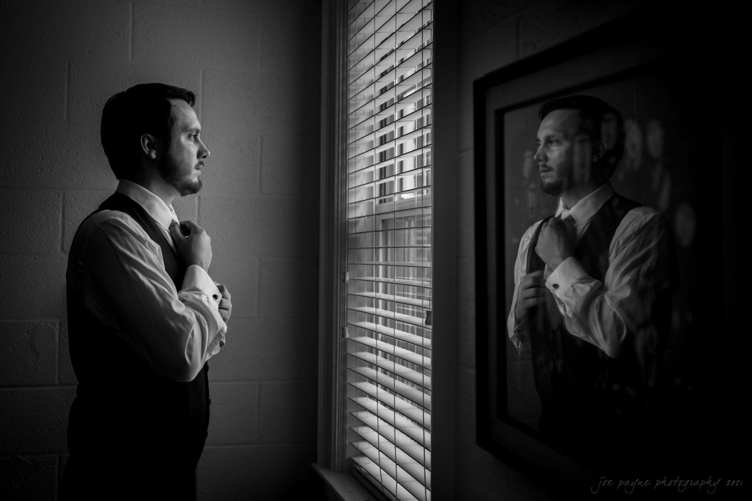 A man in formal attire stands by a window, adjusting his tie. His reflection is visible in a framed mirror across from him. The room is softly lit, creating a calm and introspective atmosphere.