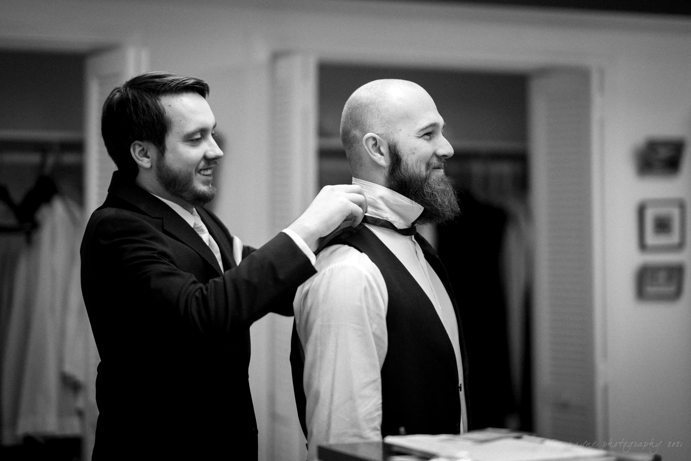 Two men in formal attire are seen in a black and white photo. One is helping the other adjust a bow tie while smiling. They stand in a room with open closets and framed pictures on the wall, creating a warm, candid atmosphere.