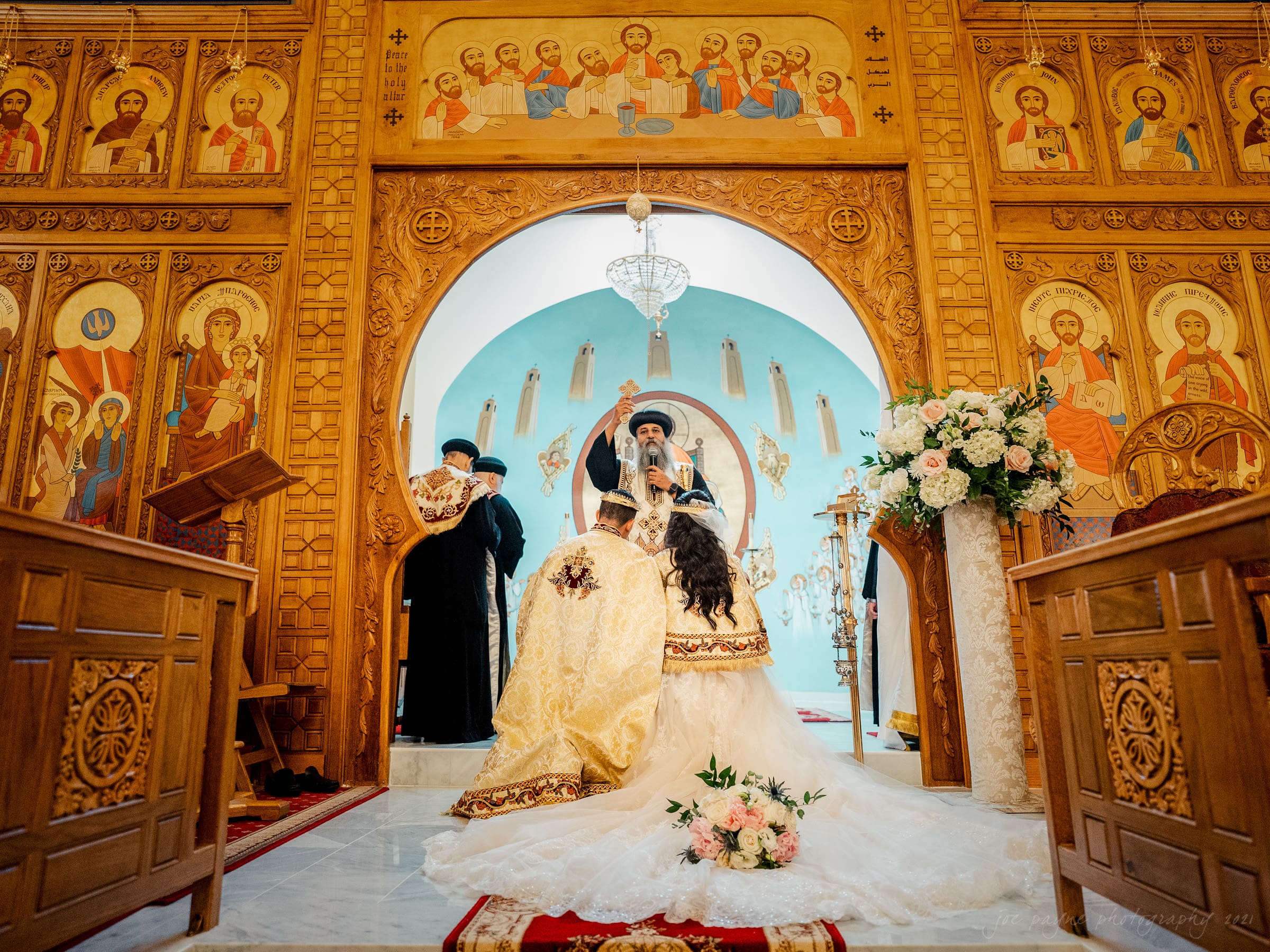 A couple in traditional attire kneels before a priest during a wedding ceremony in an ornate church. The wooden interior features religious icons, and flower arrangements adorn the space. The bride wears a white gown, and the groom wears gold vestments.