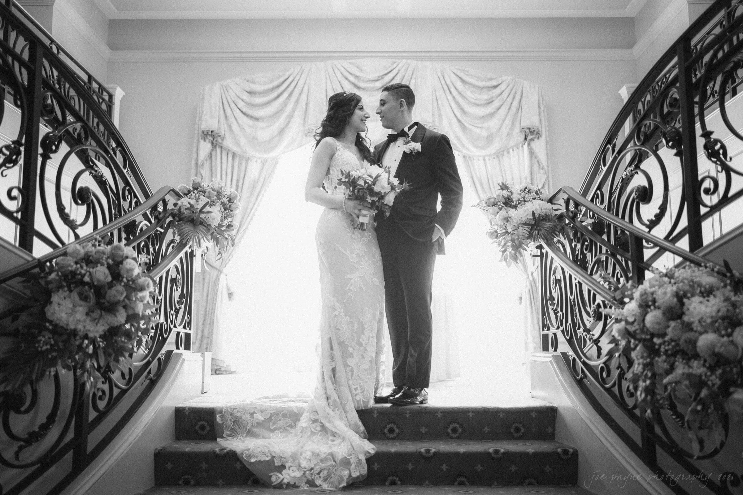 A bride and groom stand on a grand staircase, adorned with floral arrangements. The bride wears an elegant lace gown, holding a bouquet, while the groom is in a tuxedo. They are looking at each other lovingly under ornate draperies.