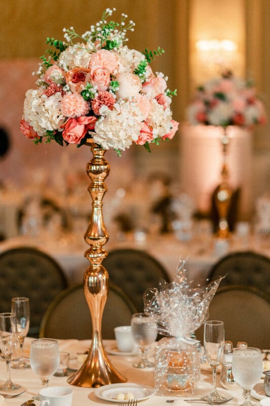 A lavish wedding table centerpiece features a tall, ornate gold stand adorned with pink and white flowers. Surrounding the centerpiece are elegant place settings with glasses and wrapped gifts on a warmly lit banquet table.