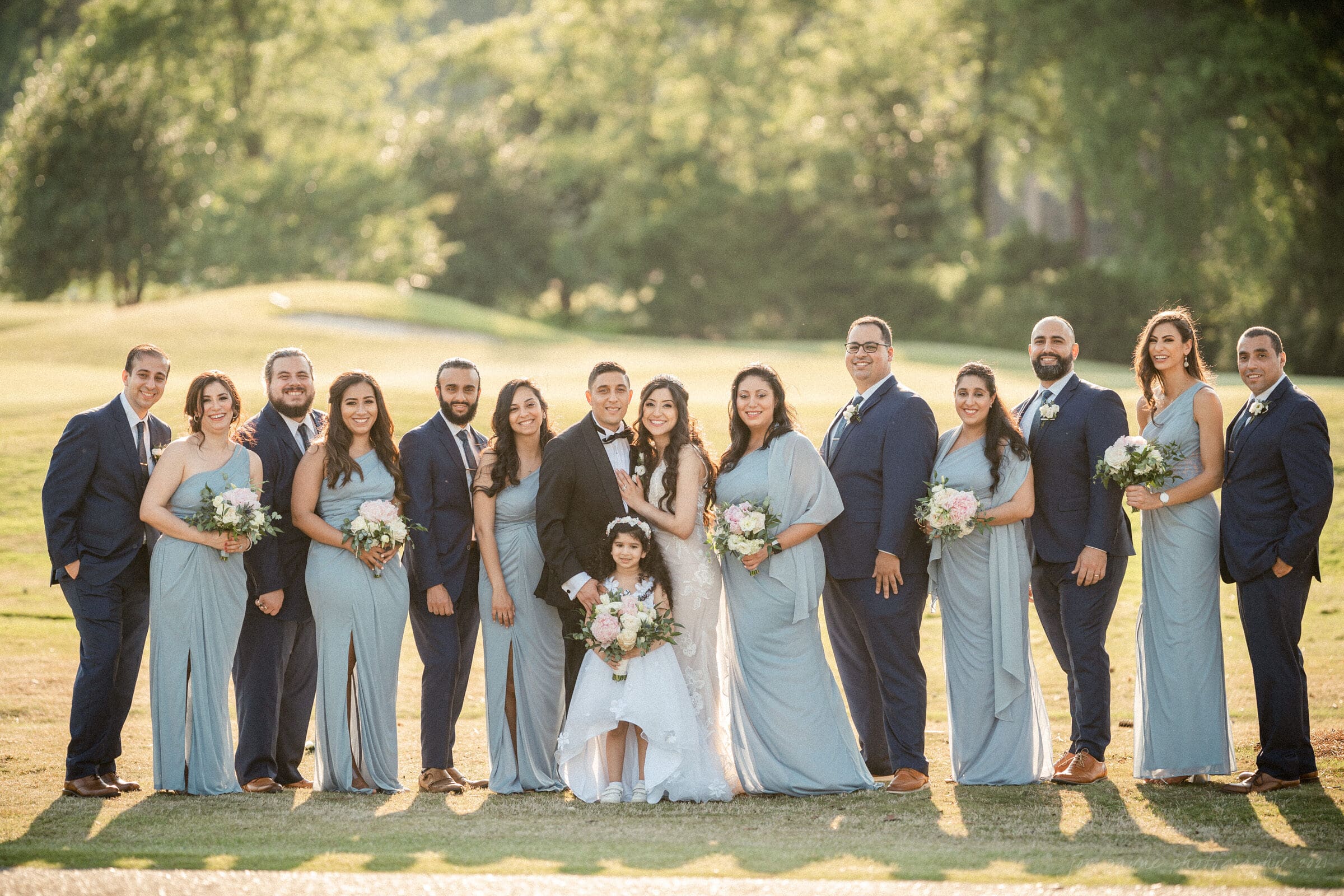A group of 15 people, including a bride, groom, and child, are posing outdoors in formal attire. The bridesmaids wear light blue dresses, and groomsmen wear navy suits. Everyone is smiling, and the background features greenery and sunlight.