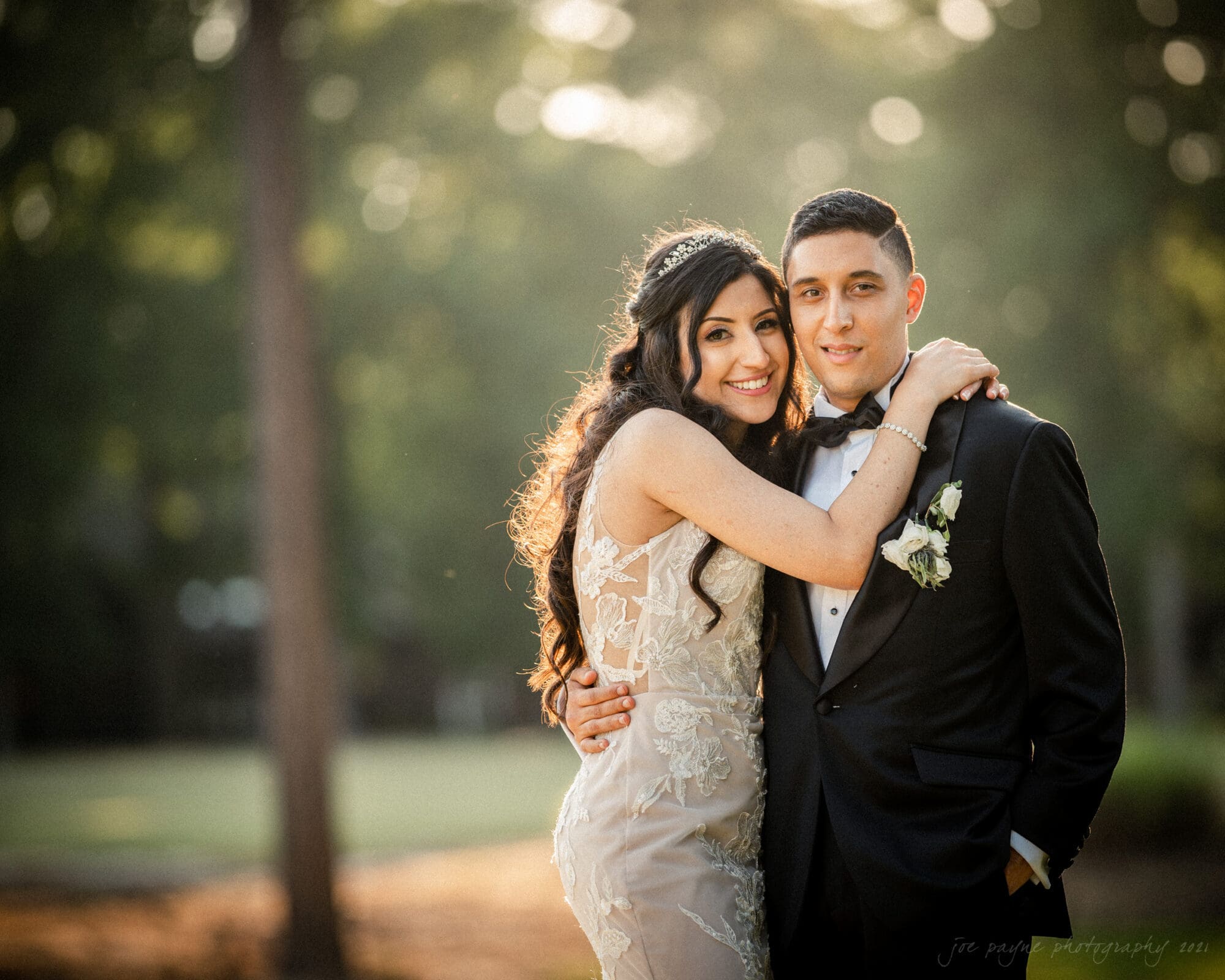 A smiling couple in formal attire stand closely together outdoors. The woman is in a lace gown and the man is in a black tuxedo with a bow tie. Sunlight filters through trees in the background, creating a soft, warm glow.