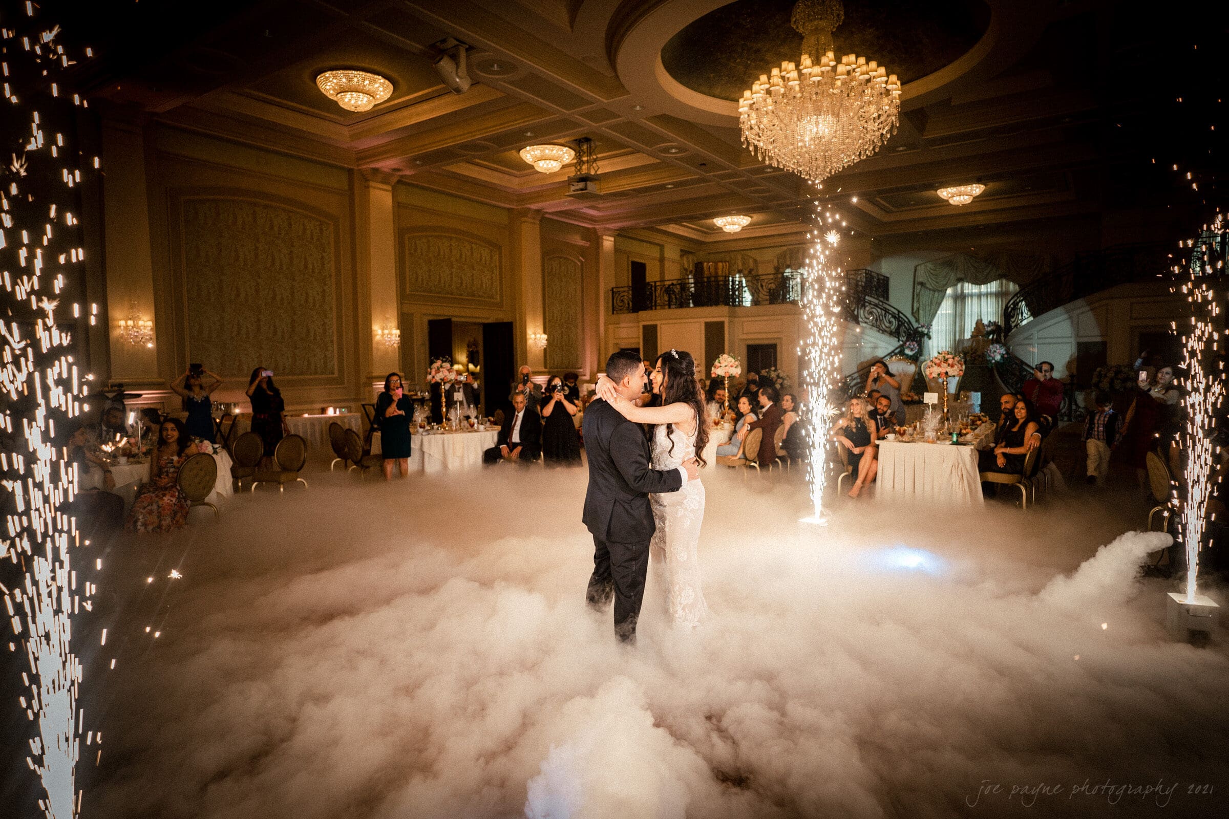 A couple dances closely in the center of a grand ballroom filled with fog and surrounded by sparkling fountain effects. Guests watch from elegantly decorated tables, and a large chandelier hangs above, casting a warm glow on the scene.