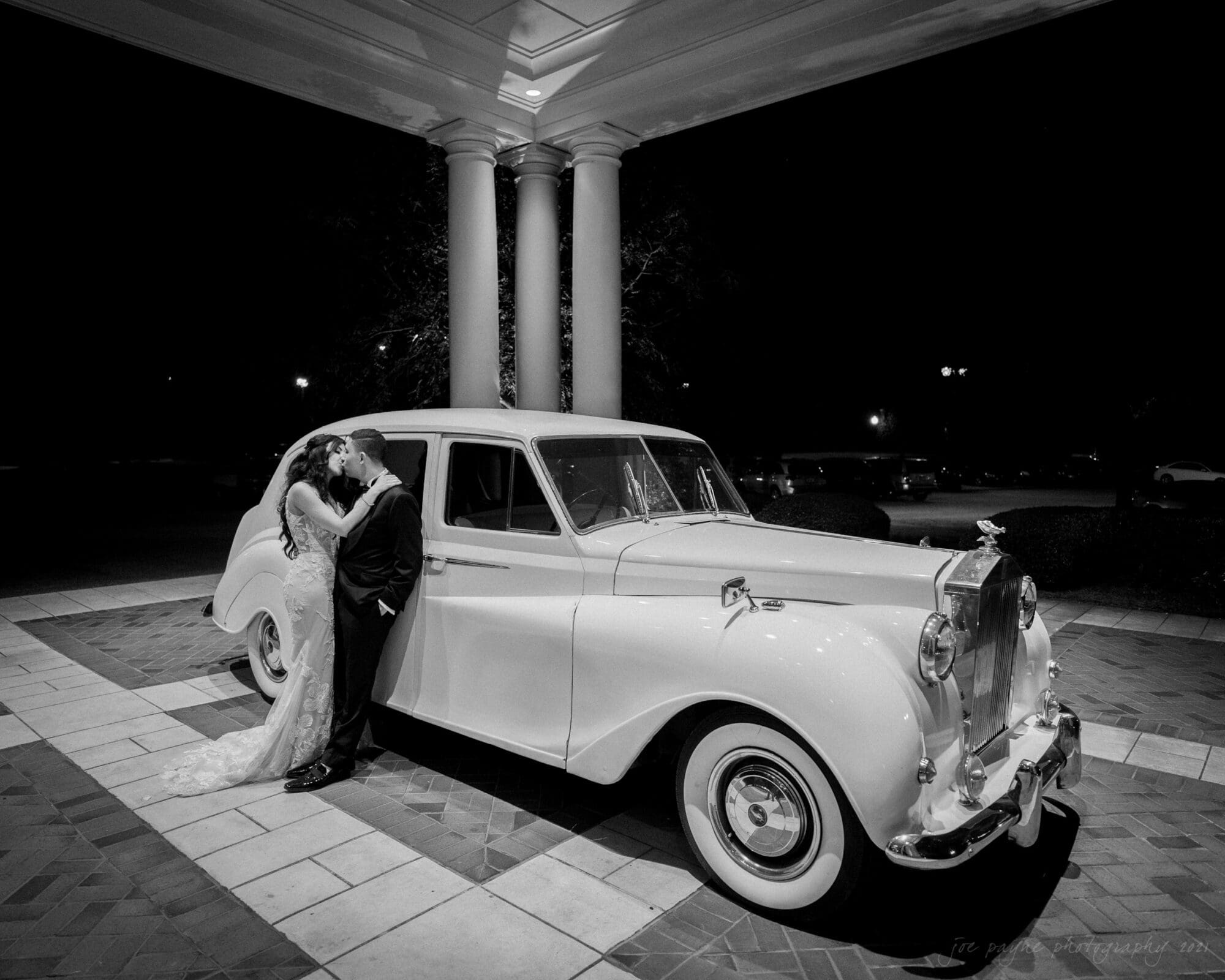 A bride and groom share a kiss beside a classic white Rolls-Royce parked under a grand portico. The bride wears a long gown with a train, and the groom is in a suit. The setting is elegant and illuminated at night.
