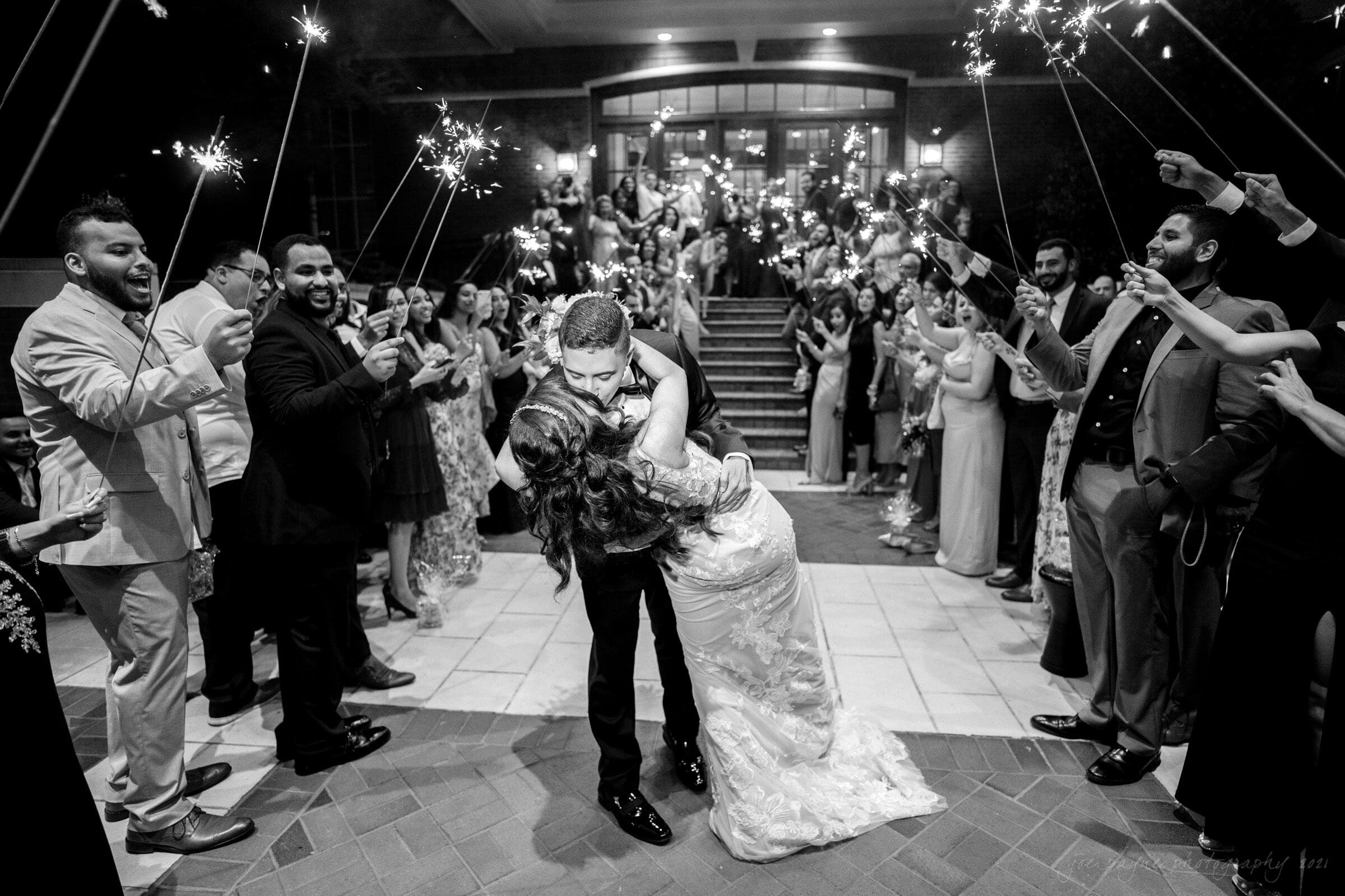 A bride and groom share a kiss while surrounded by guests holding sparklers during a nighttime celebration. The couple stands on a tiled path, and the joyful crowd forms a lively arch with their sparklers.