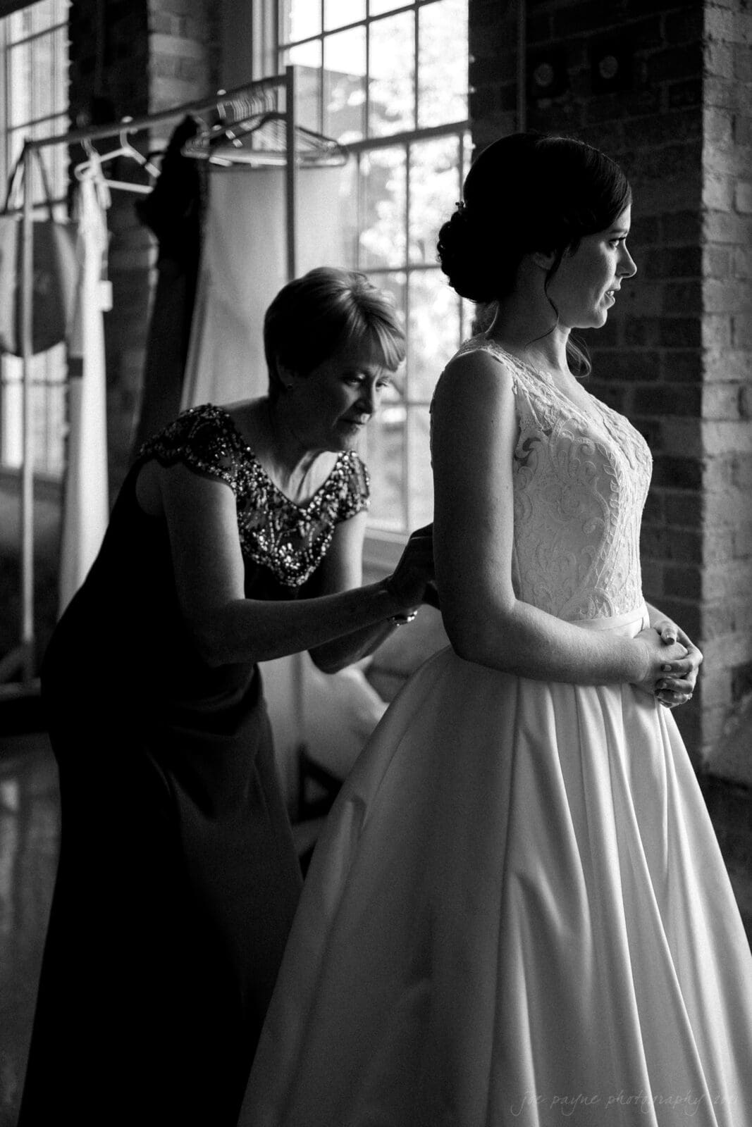 cotton room wedding photographer - kate & martin A black and white photo of a woman in a wedding dress being helped by another woman with fastening the dress. They are in a room with large windows, and several garments hang in the background.