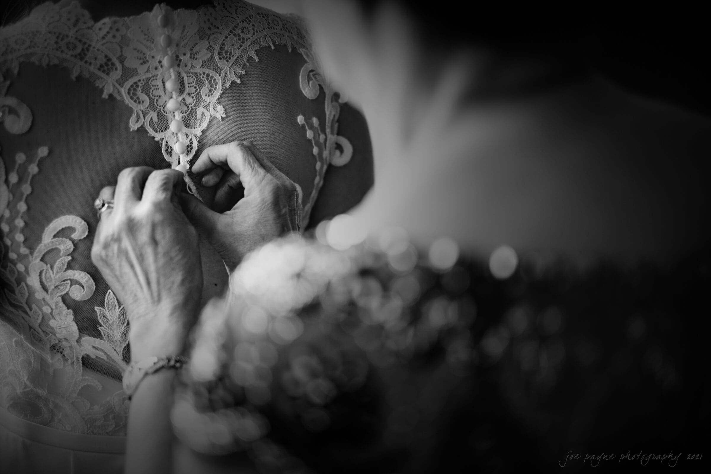 cotton room wedding photographer - kate & martin A black-and-white close-up of a person fastening buttons on the intricate lace back of a wedding dress. The focus is on the delicate embroidery and the hands, creating an intimate, elegant scene.