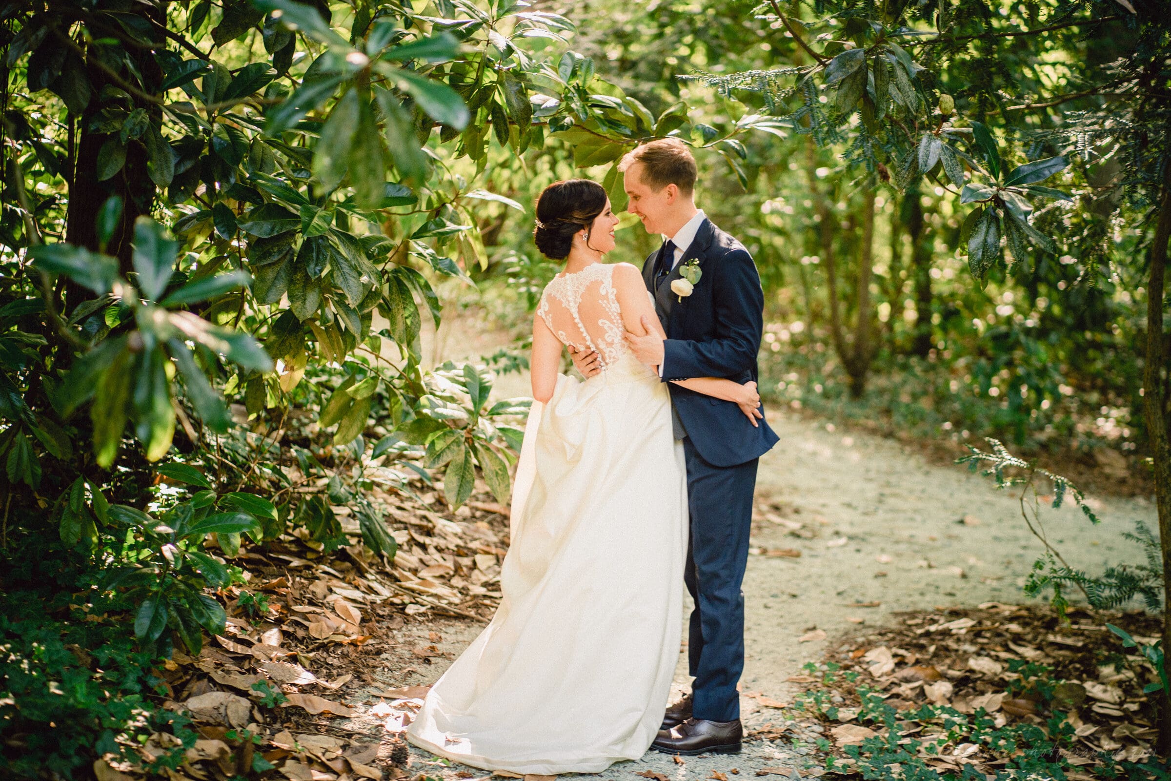 cotton room wedding photographer - kate & martin A bride and groom share an intimate moment on a sunlit forest path. She wears a white gown with a lace back, while he wears a navy suit. They are surrounded by lush green foliage, creating a romantic and serene setting.