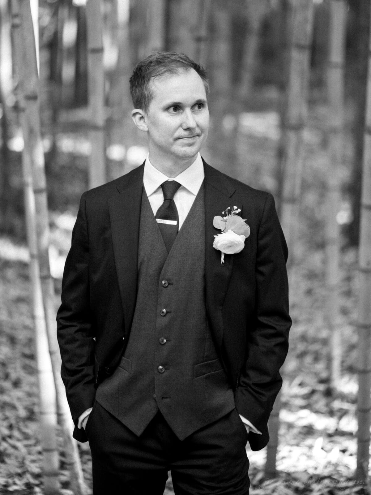 cotton room wedding photographer - kate & martin A man in a formal suit with a vest and tie stands in a bamboo forest. He wears a boutonniere and has his hands in his pockets, looking slightly to the side. The image is in black and white.
