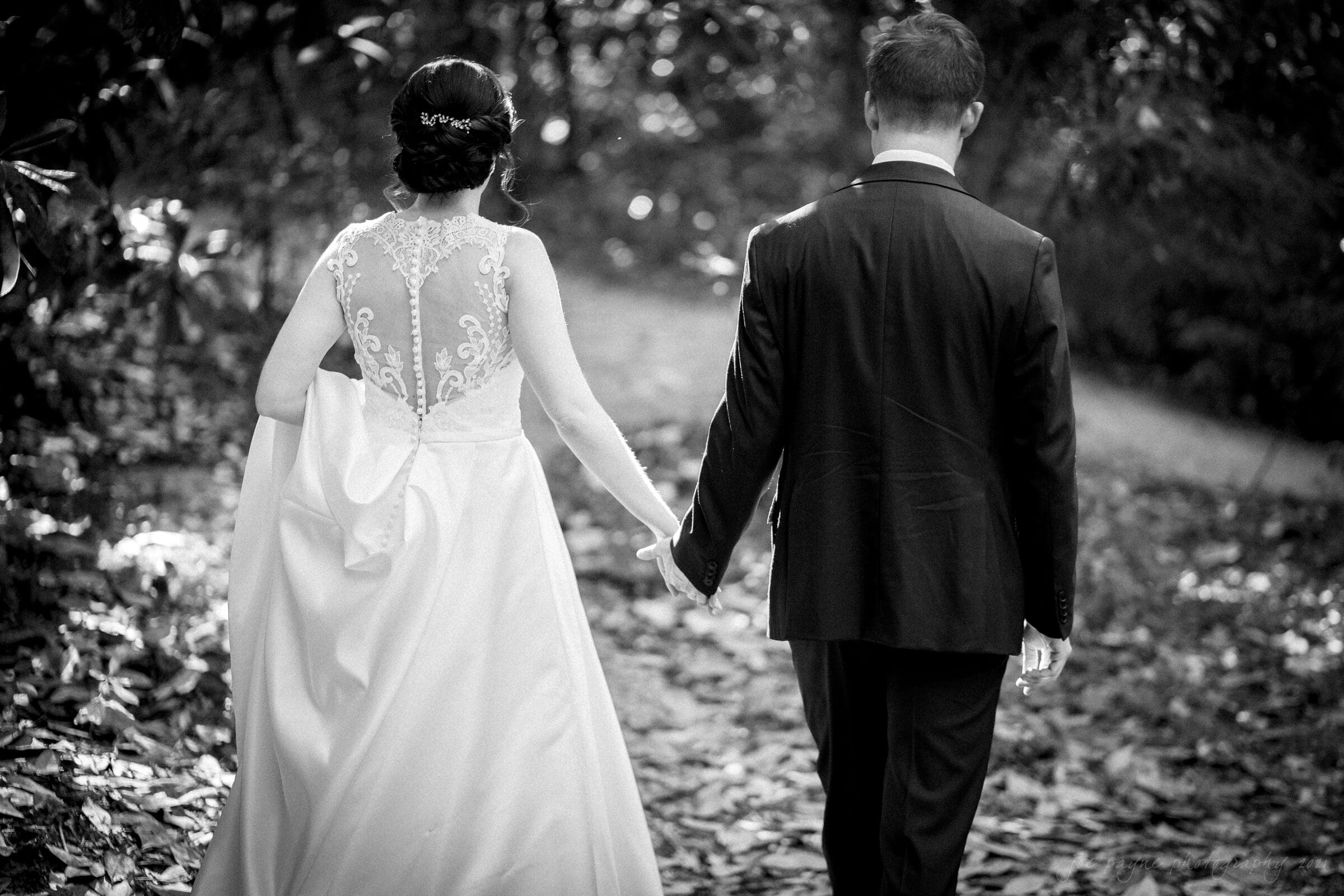 cotton room wedding photographer - kate & martin A black-and-white photo of a couple in wedding attire walking hand in hand through a wooded path. The bride wears a dress with lace details, and the groom is in a dark suit. Sunlight filters through the trees, casting soft shadows on the ground.