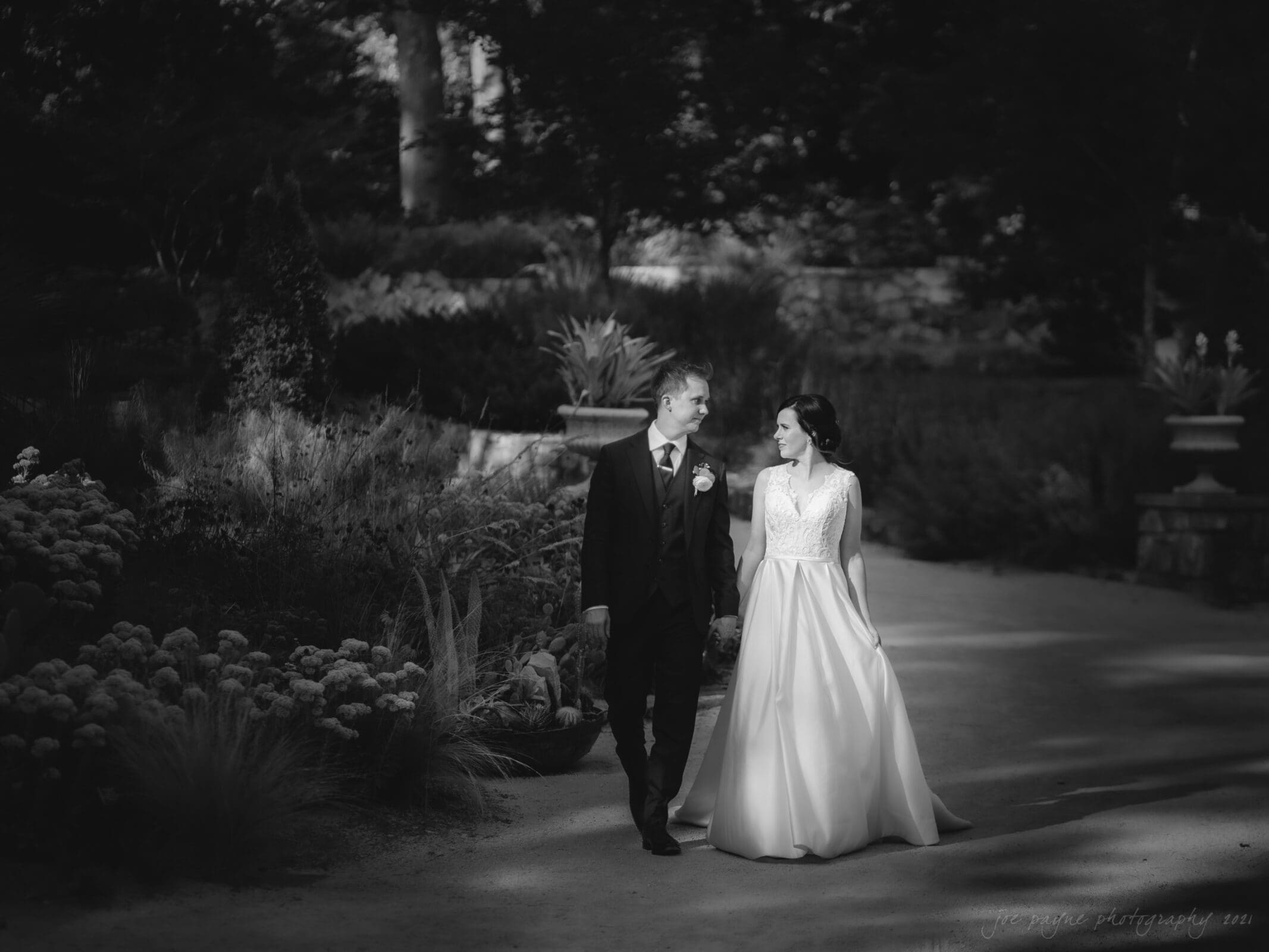 cotton room wedding photographer - kate & martin A couple in wedding attire strolls hand-in-hand through a garden. The groom wears a suit, and the bride wears a long, elegant dress. They are surrounded by foliage, illuminated gently by light filtering through the trees.