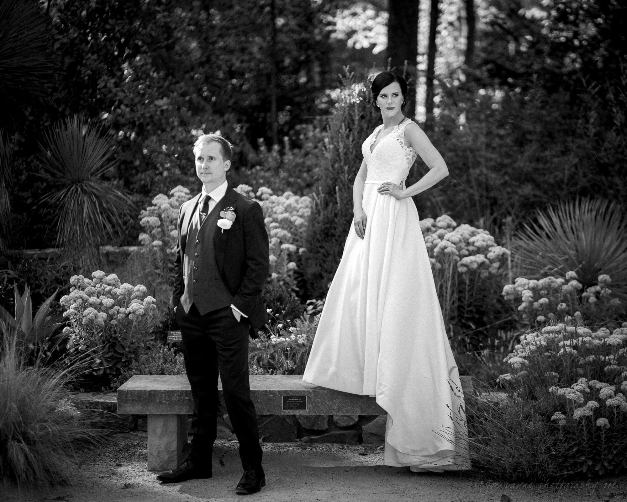 cotton room wedding photographer - kate & martin A black and white photo of a bride and groom in a garden. The bride stands confidently with one hand on her hip, wearing a flowing gown, while the groom stands beside her in a suit, hands in pockets. They are surrounded by lush foliage and flowers.