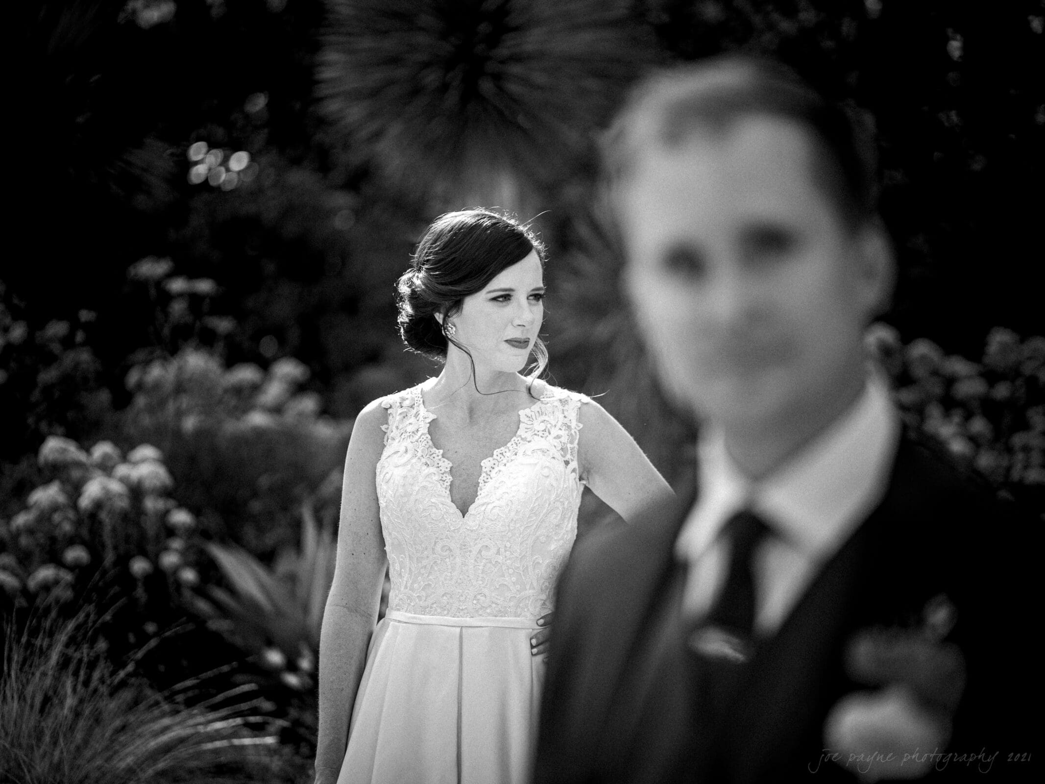 cotton room wedding photographer - kate & martin A black and white image of a bride in a lace dress looking to the side with blurred foliage in the background. A groom in focus stands in the foreground, slightly out of focus, wearing a suit.