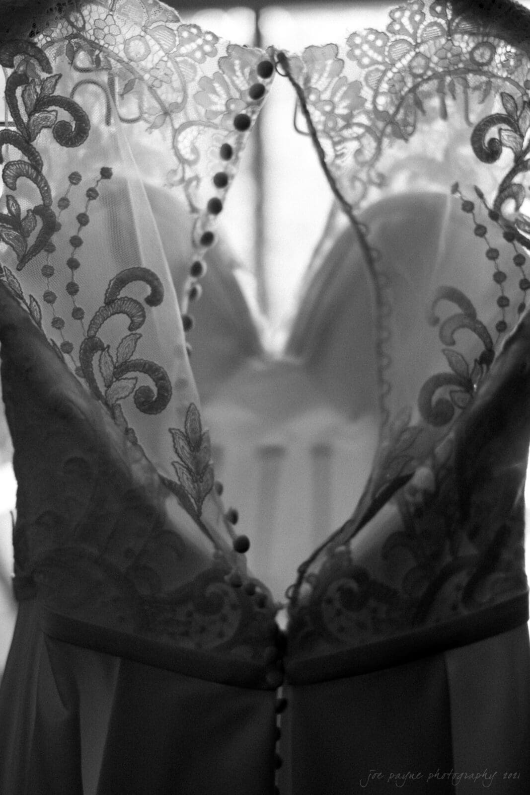 cotton room wedding photographer - kate & martin Black and white image of a wedding dress, showing intricate lace patterns and a row of buttons down the back. The dress is hanging, with soft lighting highlighting its delicate details.
