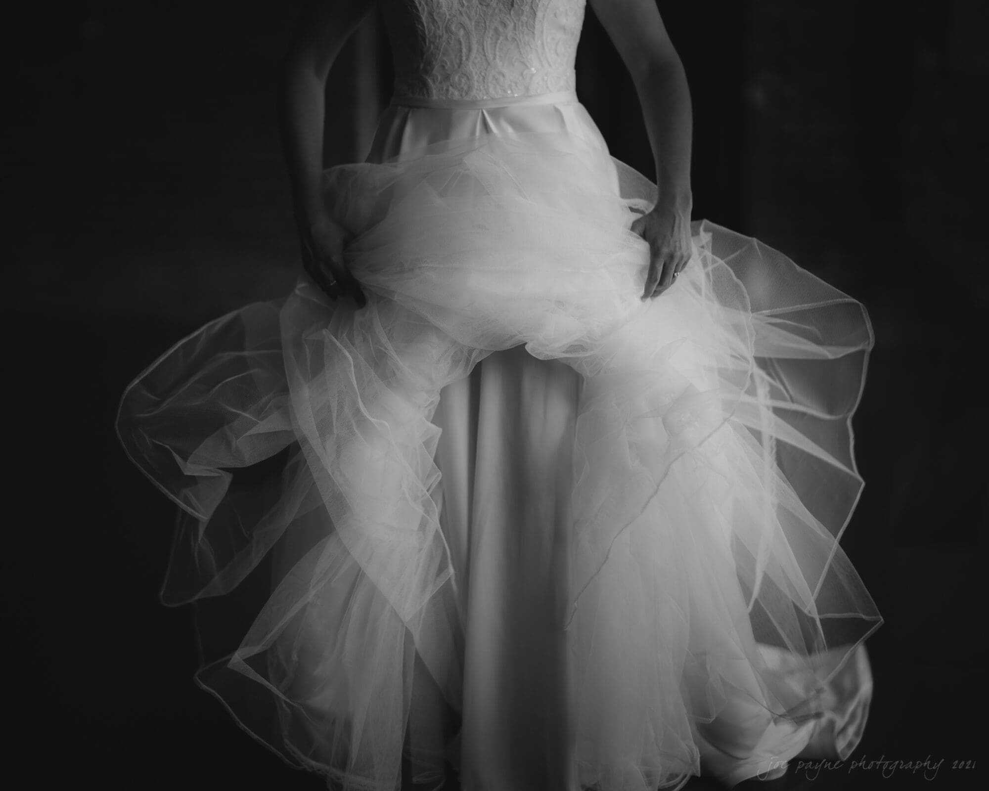 cotton room wedding photographer - kate & martin A black and white photo of a person in a wedding dress with intricate lace on the bodice, holding up the layers of the flowing tulle skirt. The image focuses on the details of the gown and the texture of the fabric.