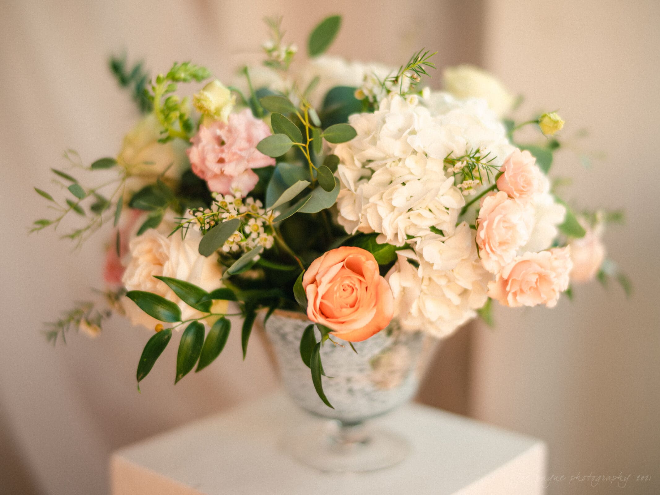 cotton room wedding photographer - kate & martin A glass vase with a floral arrangement of white hydrangeas, peach and pink roses, and green foliage. The flowers are arranged elegantly, with soft lighting highlighting their delicate petals. The background is softly blurred, adding to the serene ambiance.