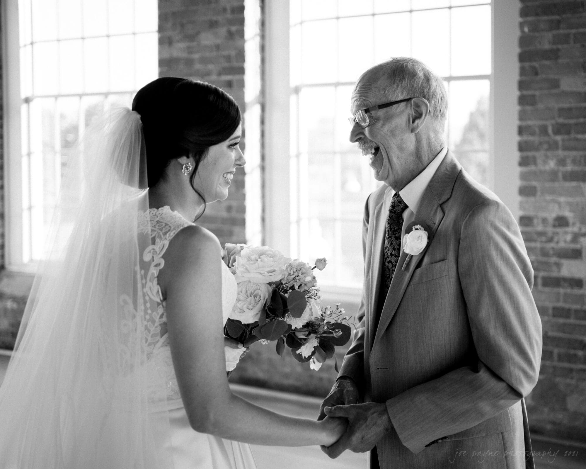 cotton room wedding photographer - kate & martin A bride in a wedding gown and veil holds a bouquet and smiles at an older man in a suit and tie. They are standing in a sunlit room with large windows and brick walls, sharing a joyful and touching moment. Black and white image.