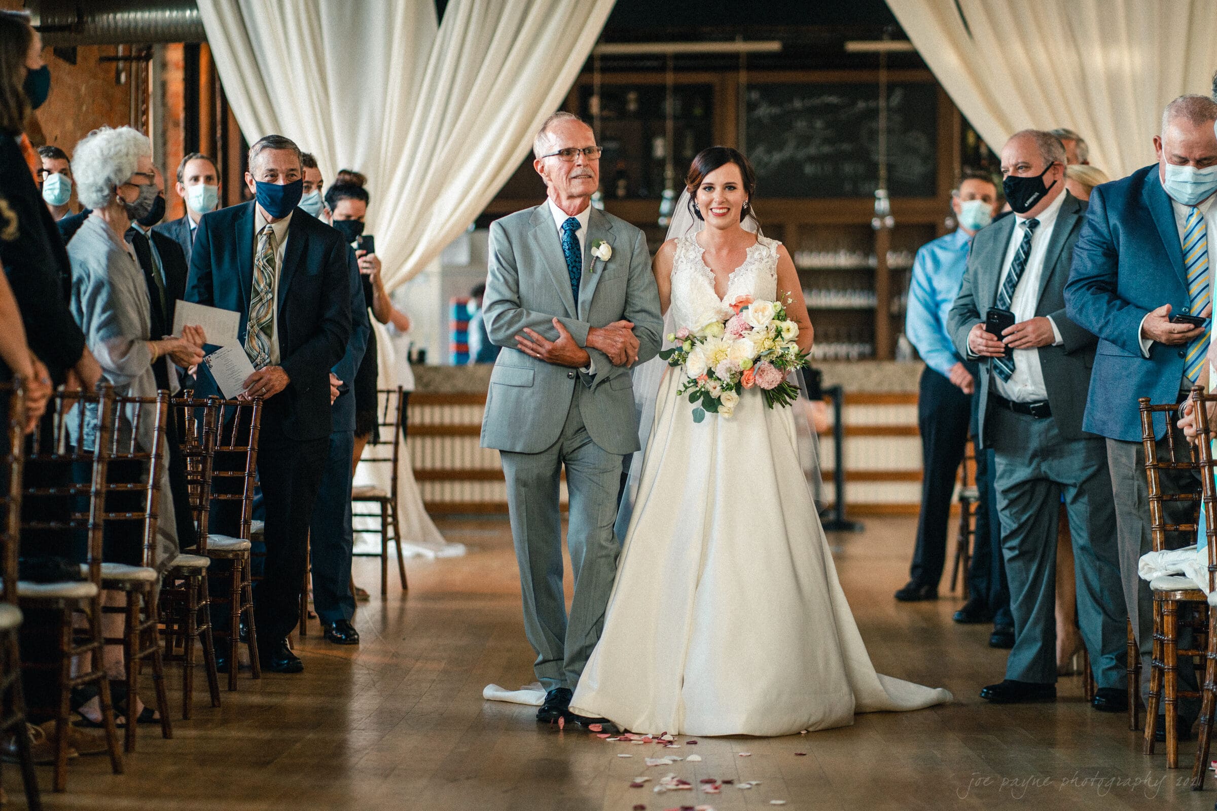 cotton room wedding photographer - kate & martin A bride in a white gown walks down the aisle with an older man in a gray suit, surrounded by seated and standing guests. She holds a bouquet of pink and white flowers. Some guests wear masks. The setting includes wooden chairs and draped curtains.