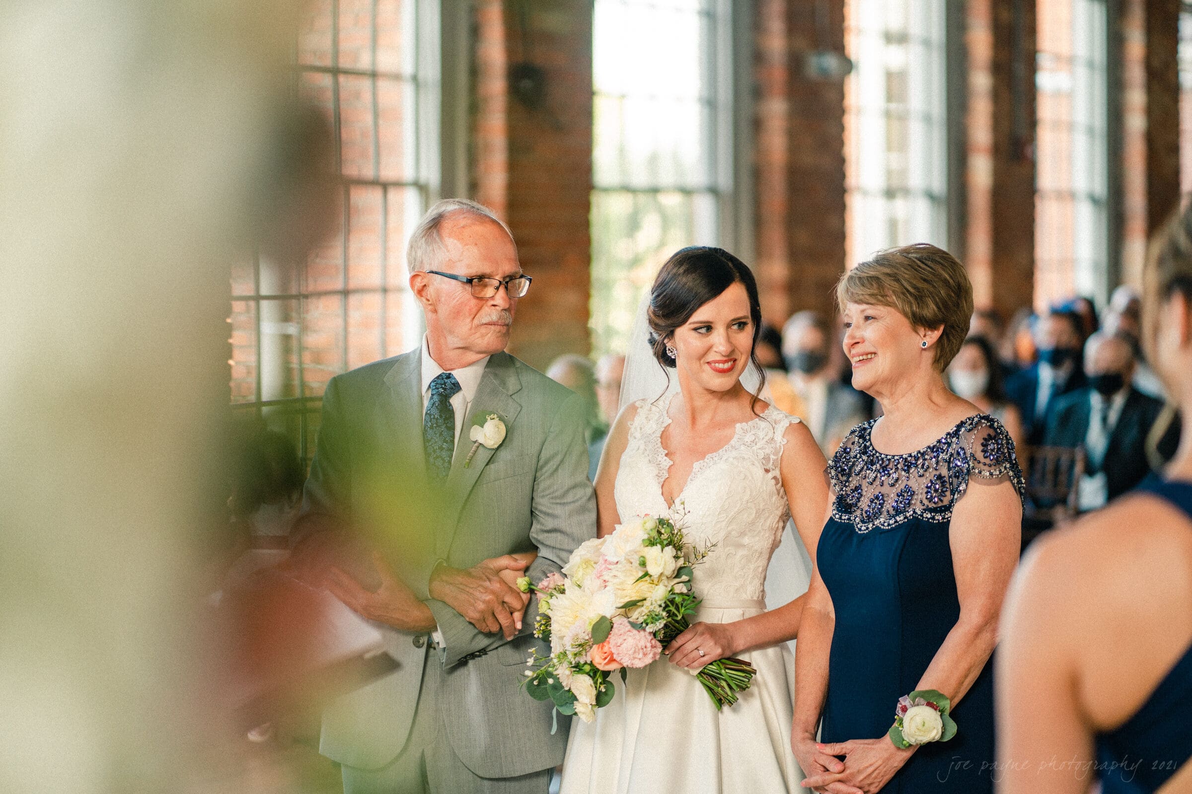 cotton room wedding photographer - kate & martin A bride in a white dress stands between two older adults, presumably her parents, in a brightly lit room. The woman on the right wears a blue dress and the man on the left is in a suit. The bride holds a bouquet of flowers.