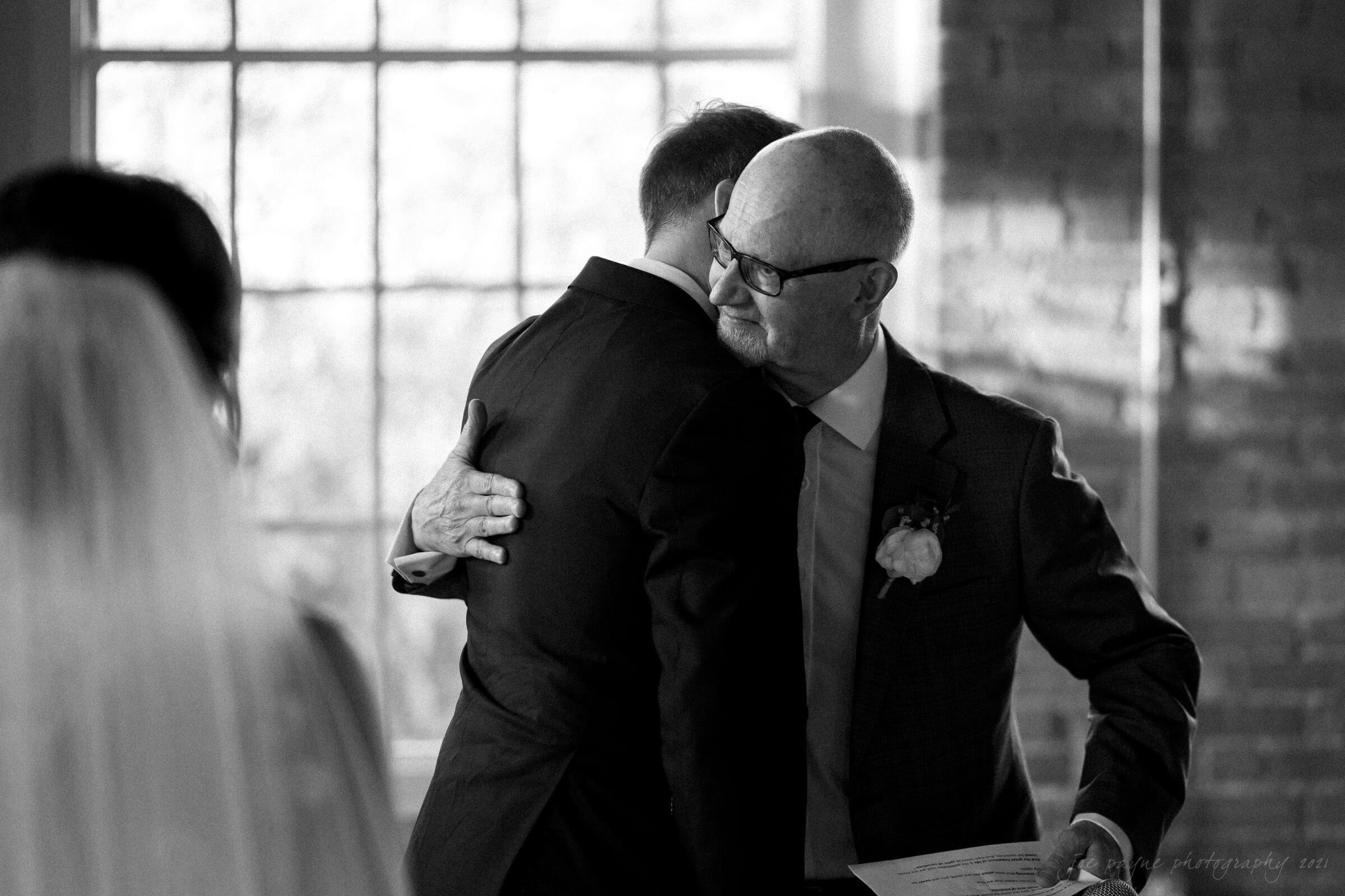 cotton room wedding photographer - kate & martin A black and white photo of an elderly man hugging a younger man in a suit, holding a piece of paper. They are in front of a large window, with a woman in a veil visible on the left. The room has brick walls and soft lighting.