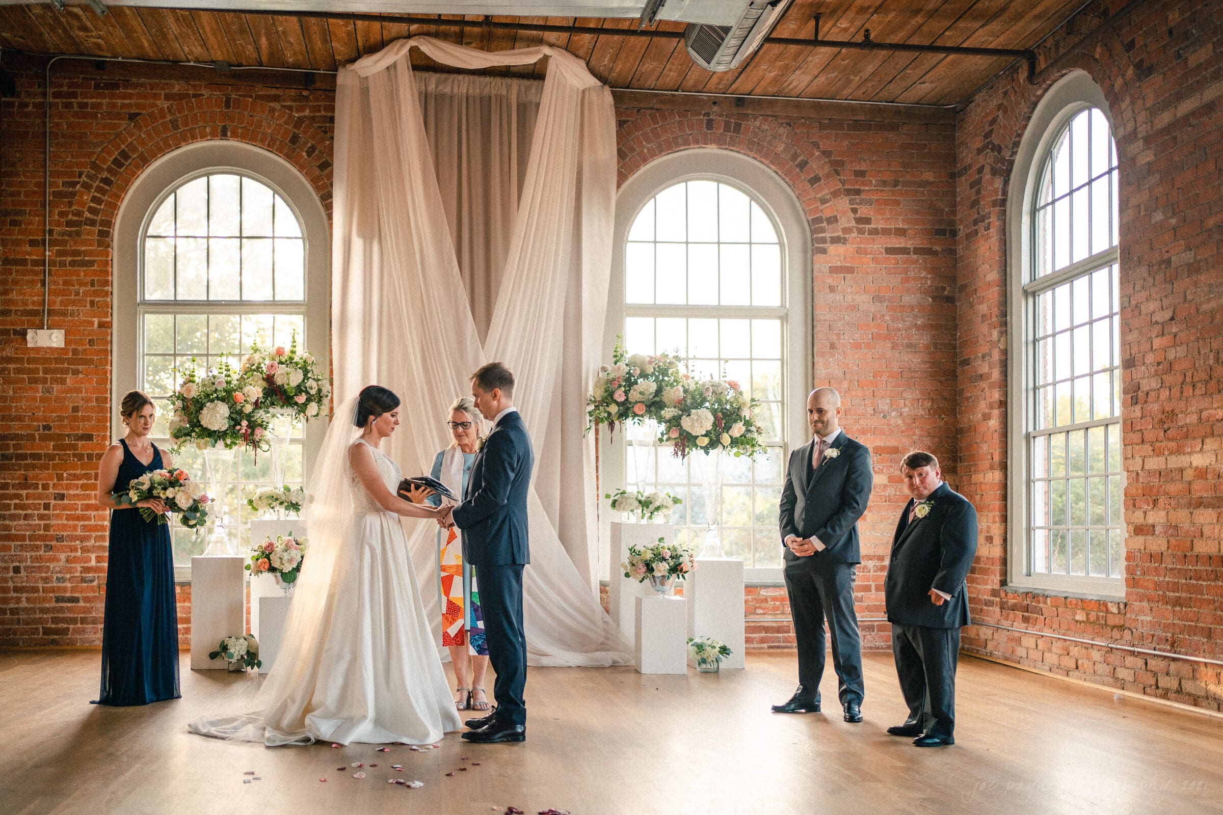 cotton room wedding photographer - kate & martin A couple stands together during a wedding ceremony in a brick-walled venue with large arched windows. The bride in a white dress and the groom in a dark suit are accompanied by an officiant. Two attendants stand on either side, surrounded by floral arrangements.