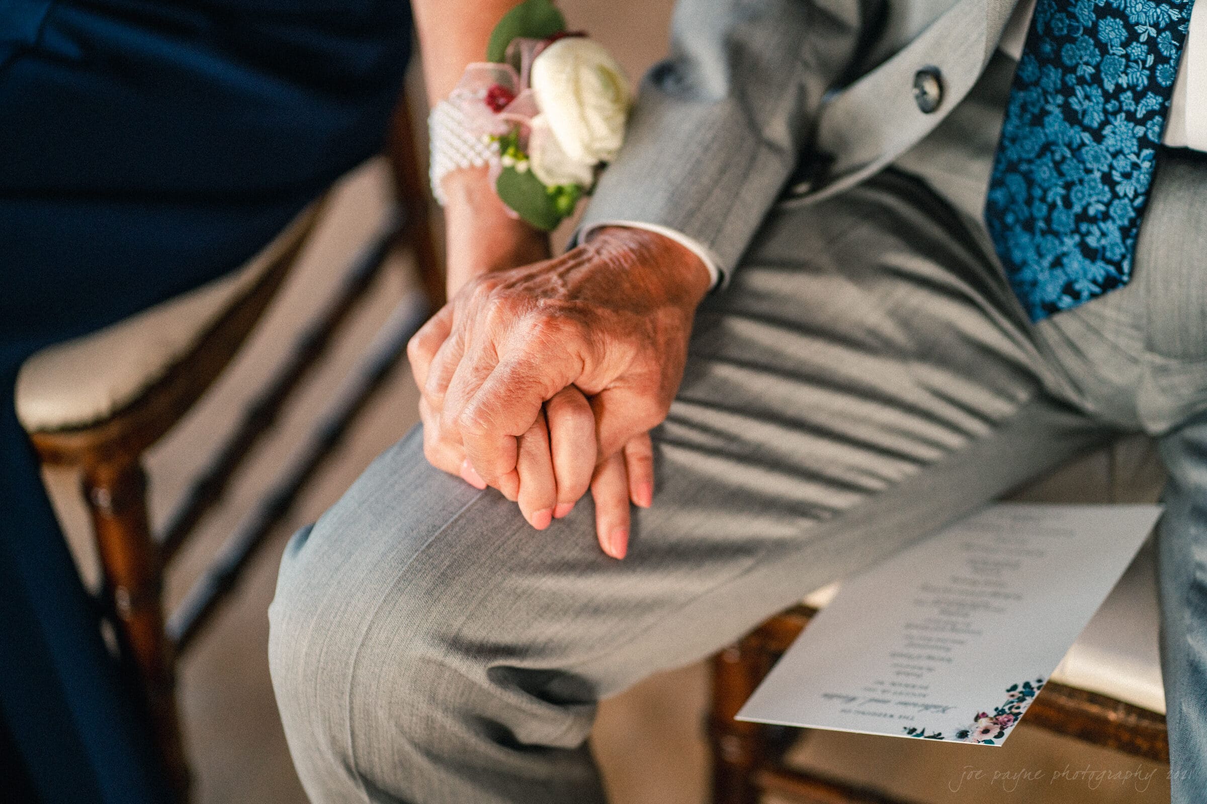 cotton room wedding photographer - kate & martin An elderly couple holding hands lovingly, seated close together. The man wears a light gray suit and floral tie, while the woman wears a corsage with a white rose. A printed paper rests on his leg.