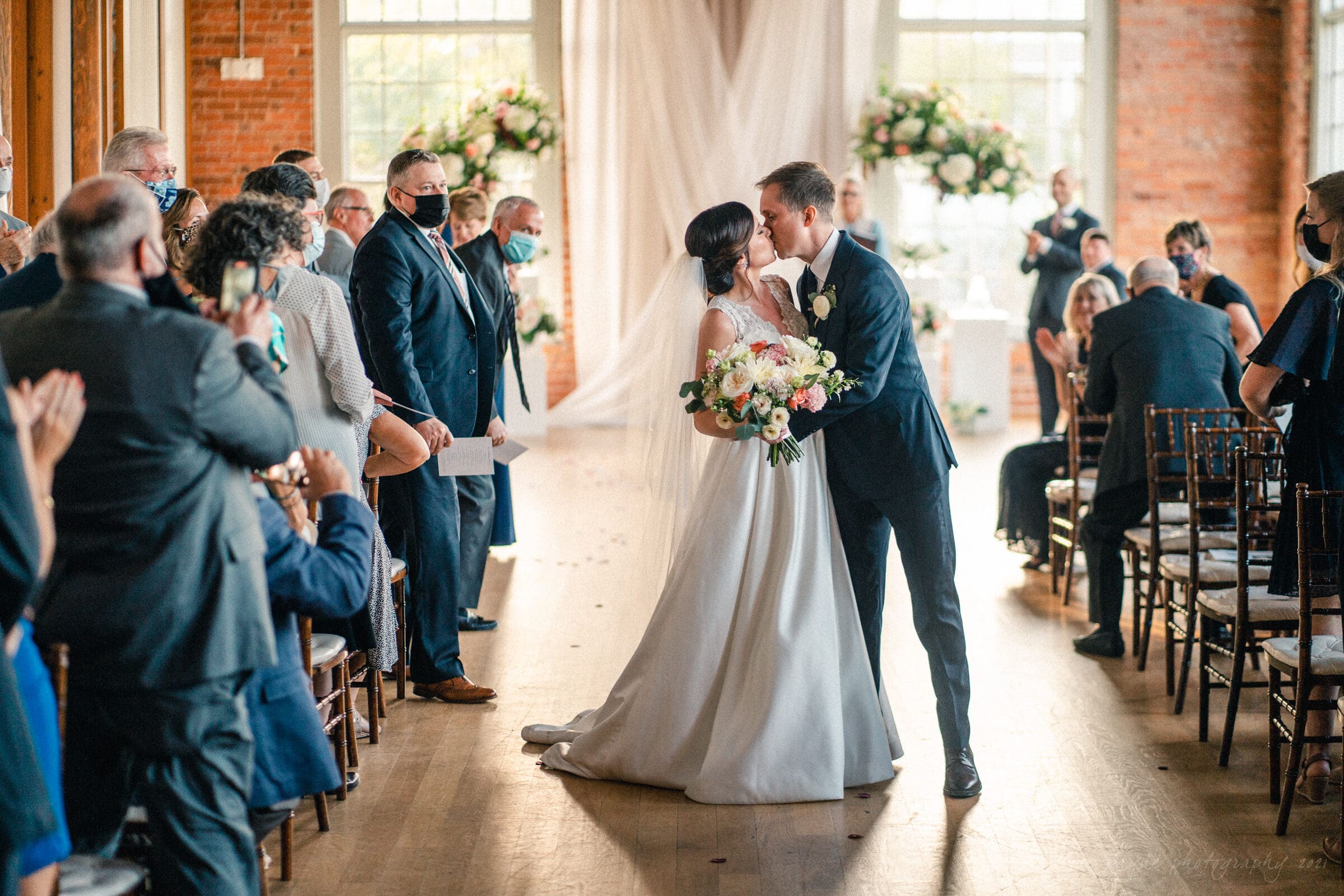 cotton room wedding photographer - kate & martin A newlywed couple shares a kiss while holding a bouquet, walking down an aisle flanked by applauding guests. The ceremony is indoors with brick walls and floral arrangements in the background. Guests wear formal attire, some with masks.