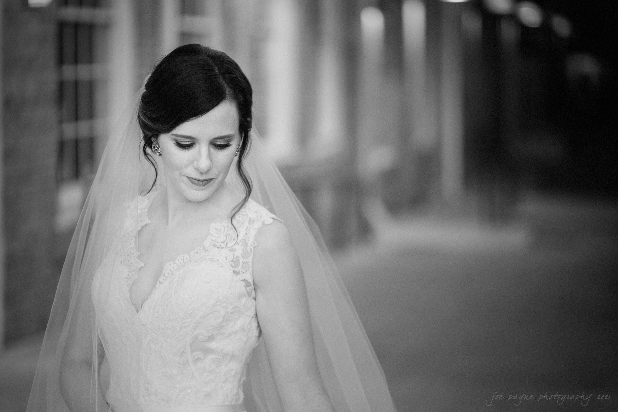 cotton room wedding photographer - kate & martin Black and white photo of a bride in a lace wedding dress with a veil. She is looking down slightly to the side, standing in a softly lit corridor or hallway.