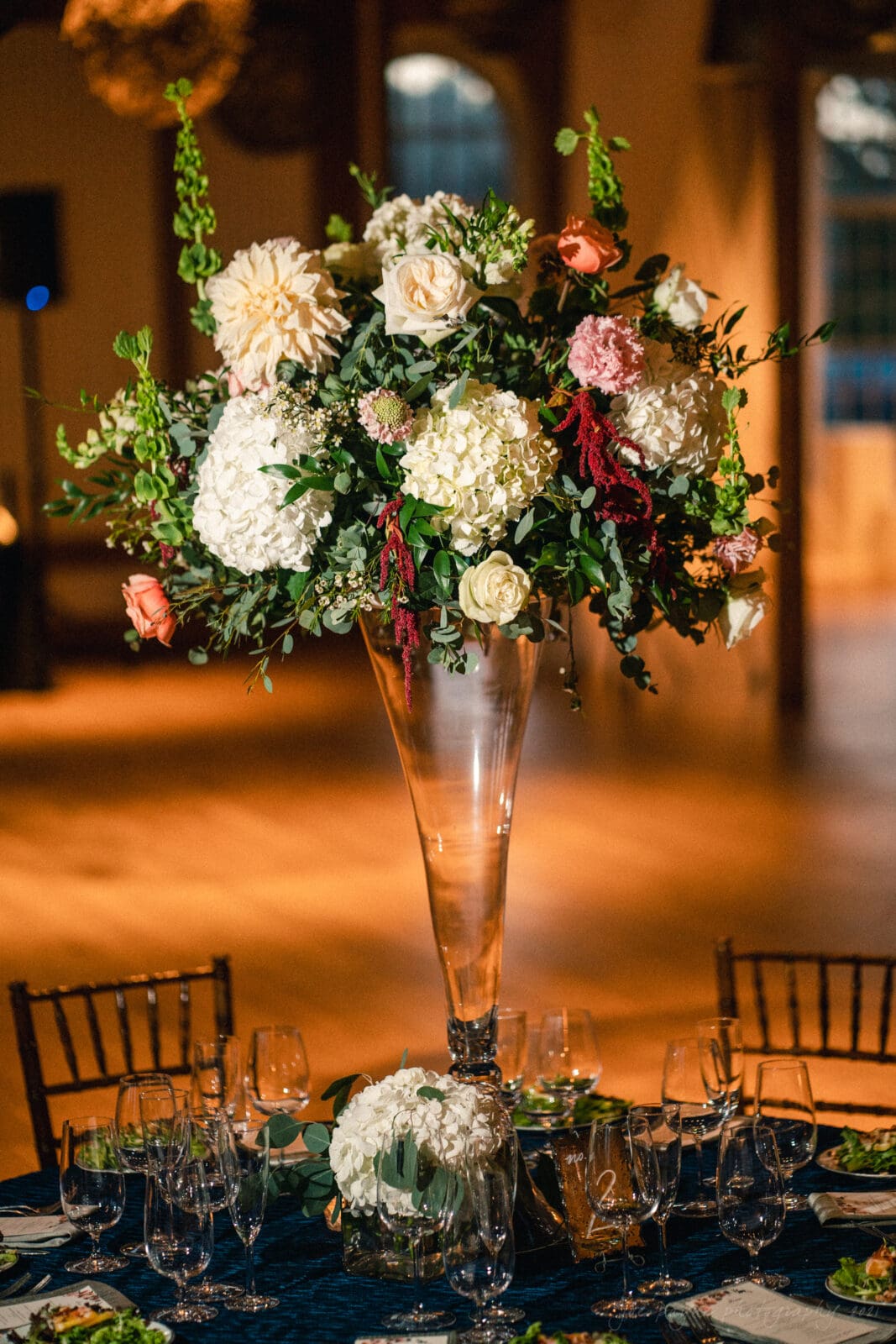 cotton room wedding photographer - kate & martin A tall, elegant floral centerpiece with white, peach, and pink flowers, including roses and hydrangeas, in a clear glass vase. Surrounded by wine glasses and wooden chairs on a dimly lit table setting.