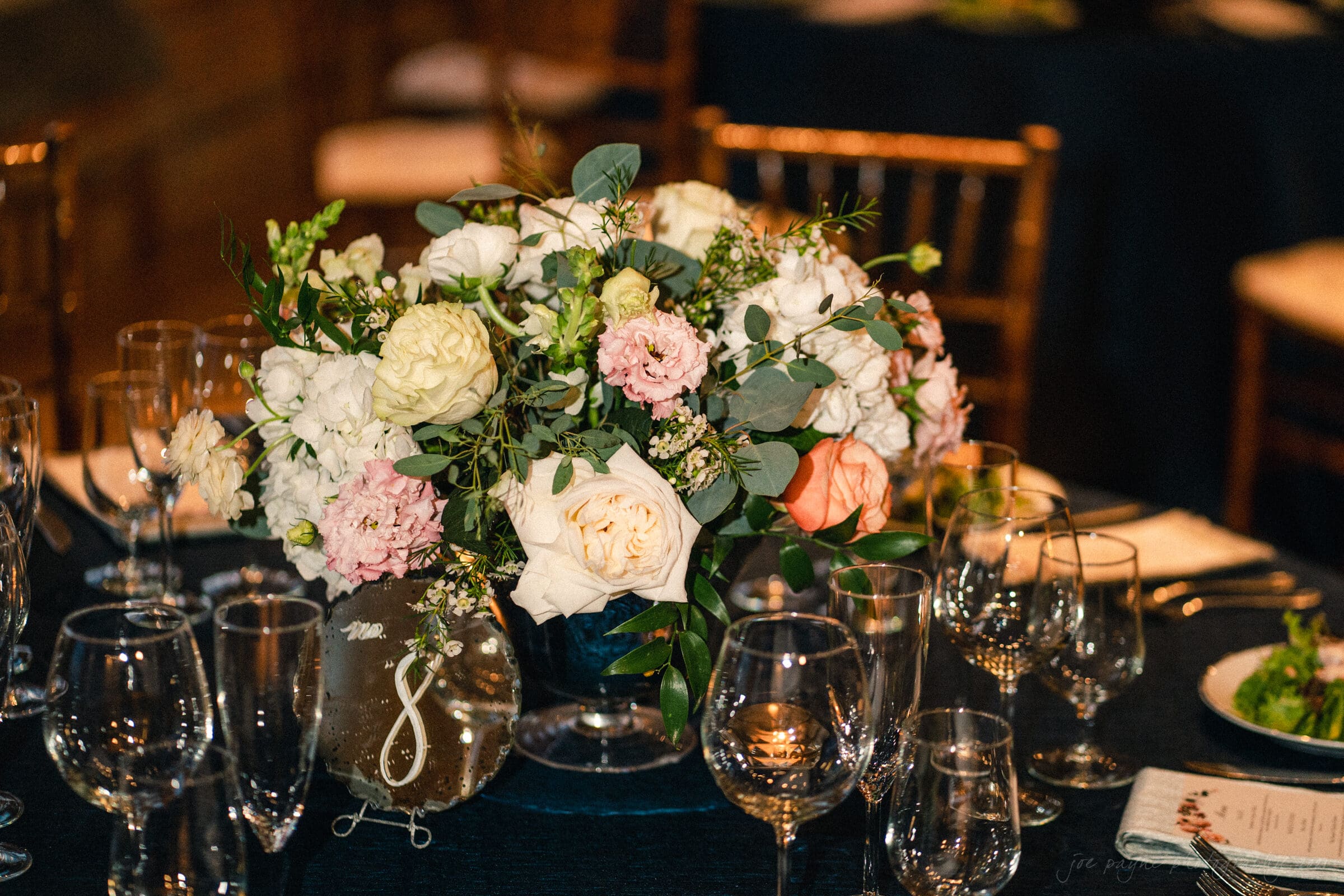 cotton room wedding photographer - kate & martin A beautifully arranged floral centerpiece with white, pink, and peach roses, surrounded by greenery on a dining table. The table is set with empty wine glasses, plates, and silverware, creating an elegant and formal atmosphere.