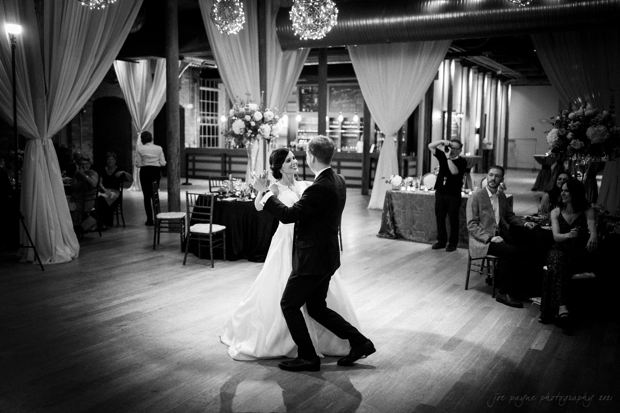 cotton room wedding photographer - kate & martin A bride and groom share a first dance in a large, elegantly decorated venue. They are surrounded by guests seated at tables with floral arrangements. The room is adorned with draped curtains and hanging lights.