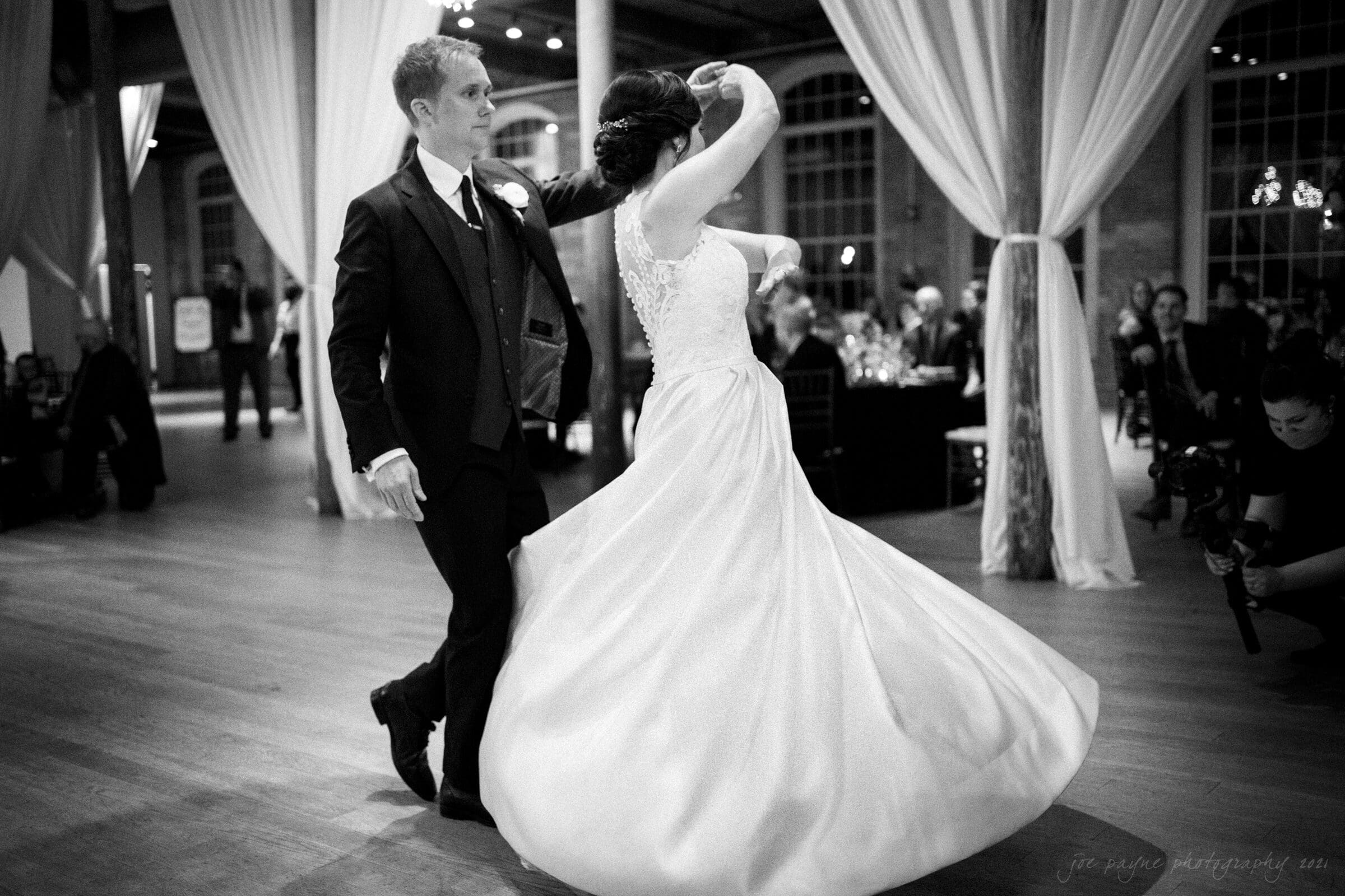cotton room wedding photographer - kate & martin A black and white photo of a wedding dance. A groom in a suit twirls the bride in a flowing gown. They are on a wooden floor, with large windows and draped curtains in the background. Guests sitting at tables watch them.
