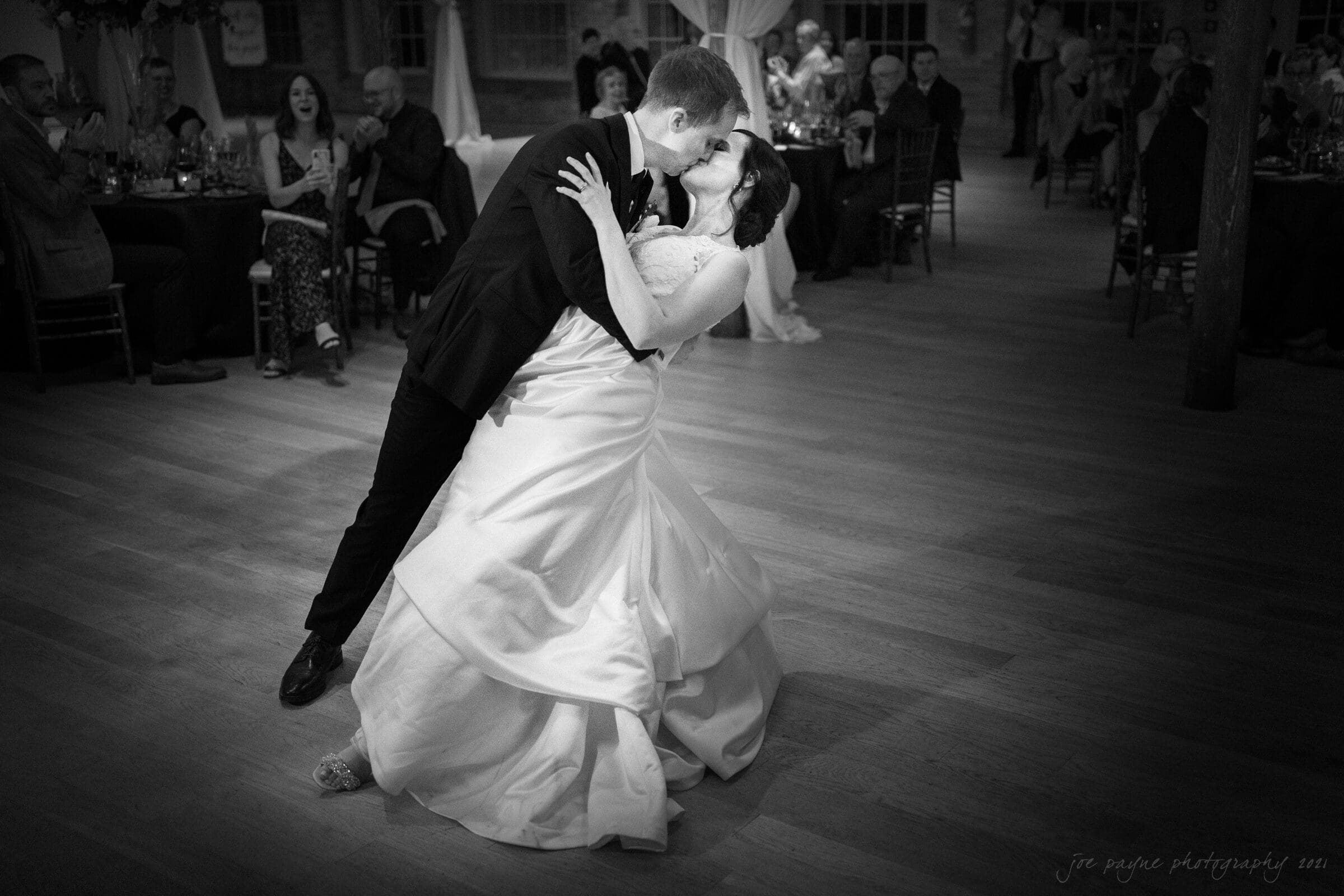 cotton room wedding photographer - kate & martin A couple shares a romantic dance on a wooden floor, with the groom dipping the bride in a graceful embrace. Guests at round tables look on, capturing the joyful and elegant atmosphere of a wedding celebration.