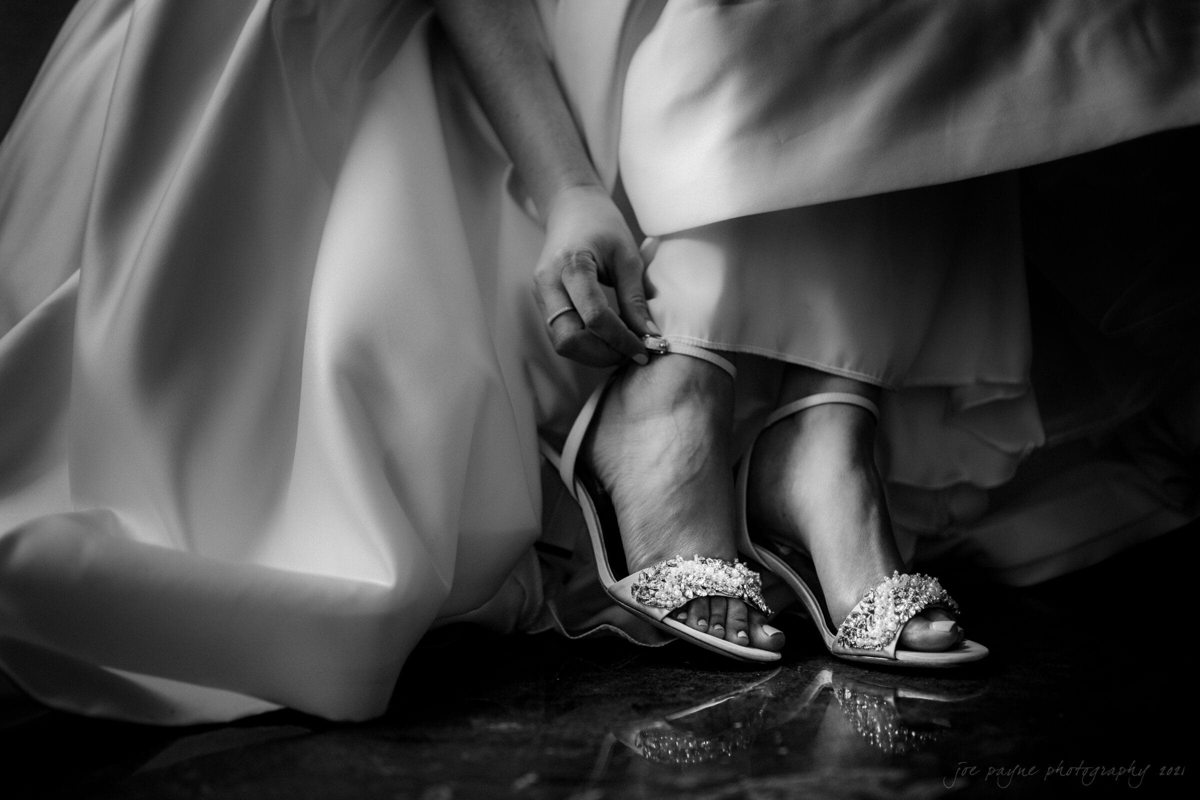 cotton room wedding photographer - kate & martin A black and white photo of a person wearing a flowing gown, adjusting an elegant strappy sandal adorned with decorative elements. Their feet are partially visible beneath the fabric. The scene exudes a sense of preparation and elegance.