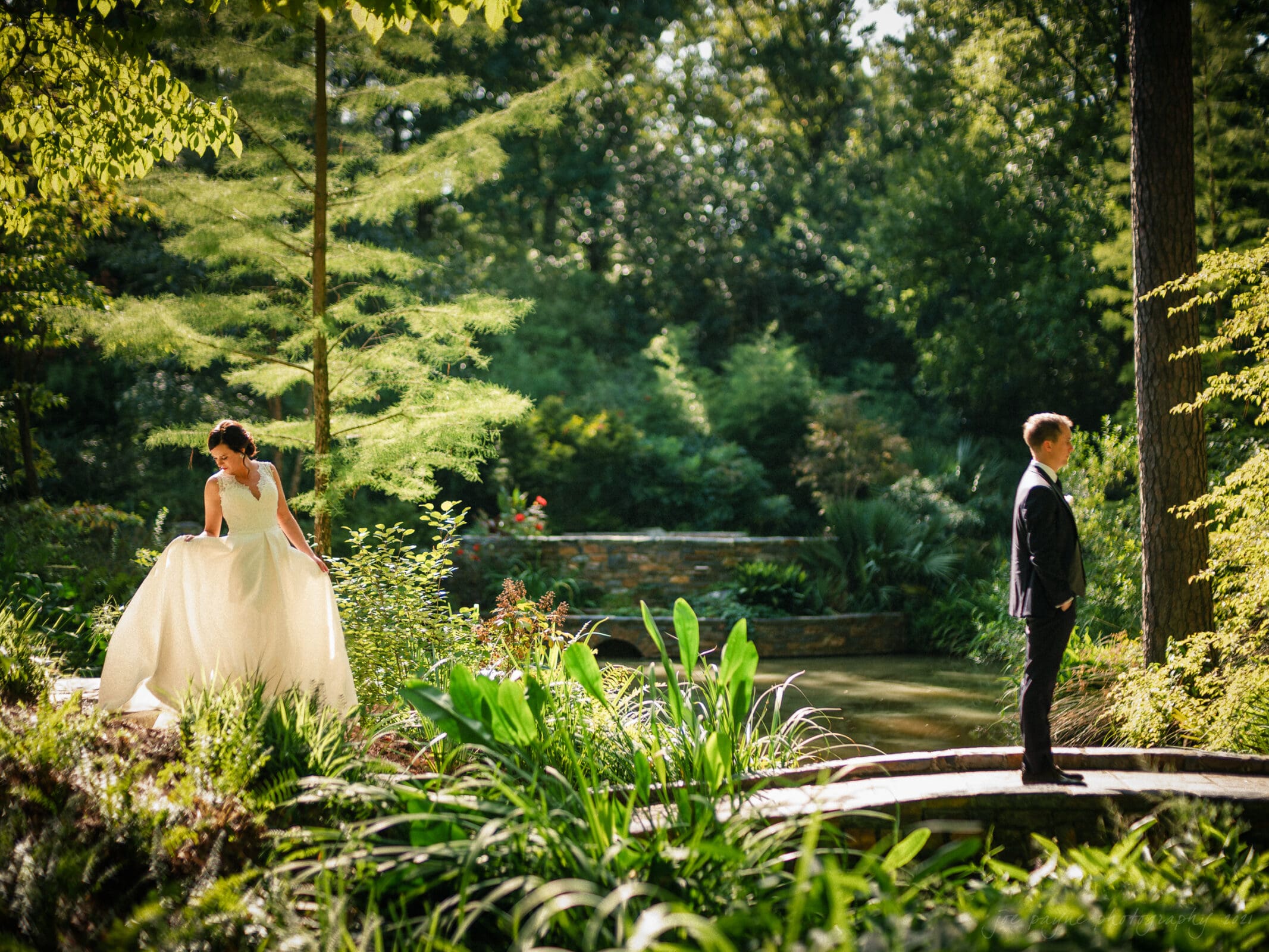 cotton room wedding photographer - kate & martin A bride in a flowing white dress stands on a stone path among lush greenery, looking at the groom who stands facing away in a black suit. Sunlight filters through the trees, creating a serene, scenic backdrop.