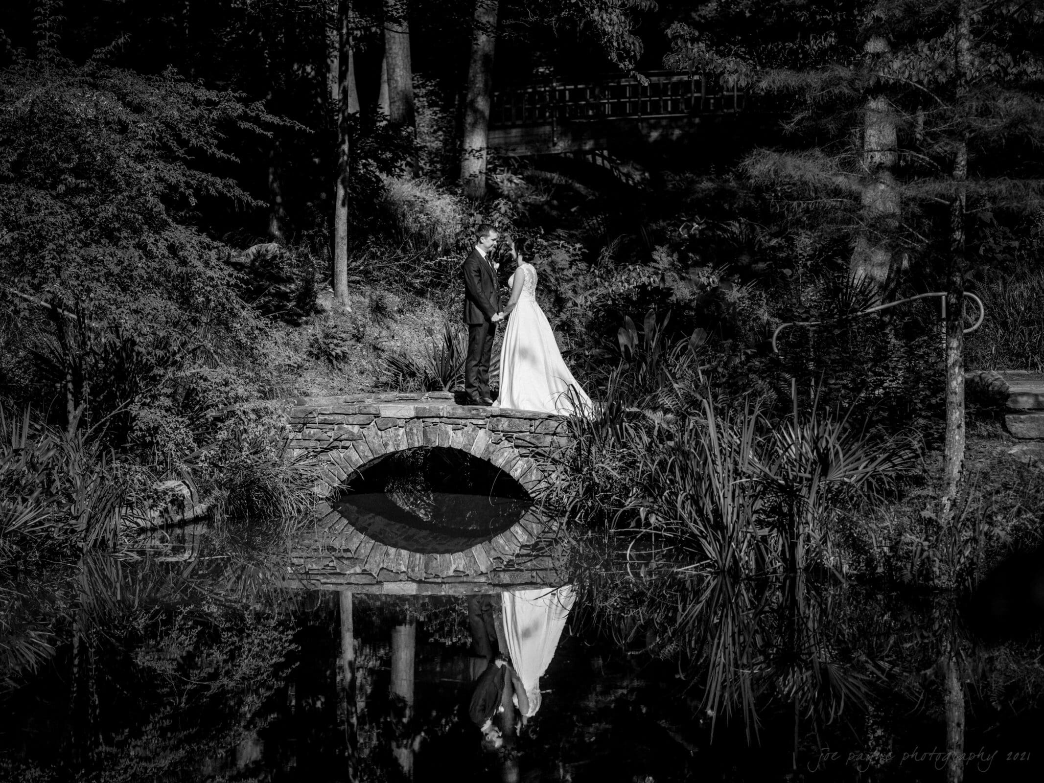 cotton room wedding photographer - kate & martin A bride and groom stand on a small stone bridge surrounded by lush foliage. The couples reflection is visible in the calm water below. The image is in black and white, adding a timeless, romantic feel to the scene.