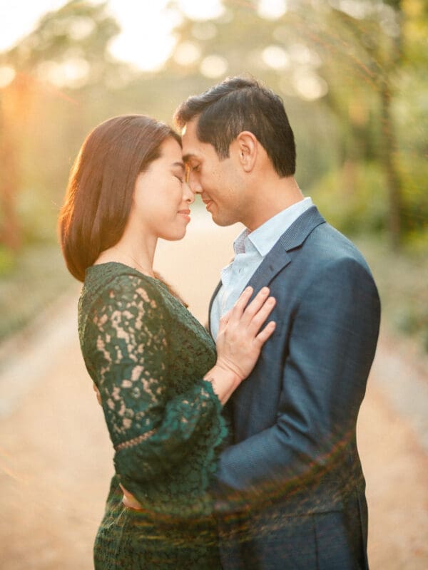 A couple gently touches foreheads, embracing on a sunlit path. The woman wears a dark green lace dress, and the man is in a blue suit. Soft dappled sunlight filters through the trees, creating a warm, romantic atmosphere.
