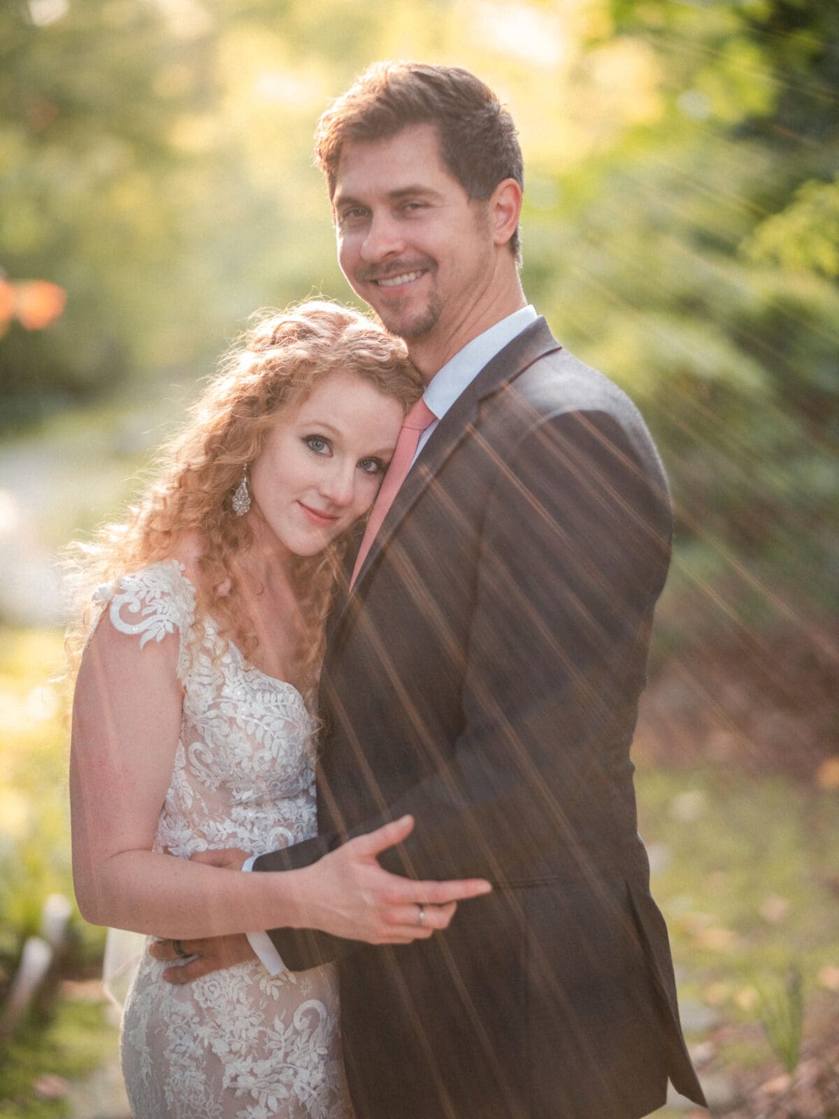 A couple in a forest setting embraces warmly under sunlit rays. The woman wears a lace wedding dress, and the man is in a suit with a pink tie. They both smile, surrounded by lush greenery.