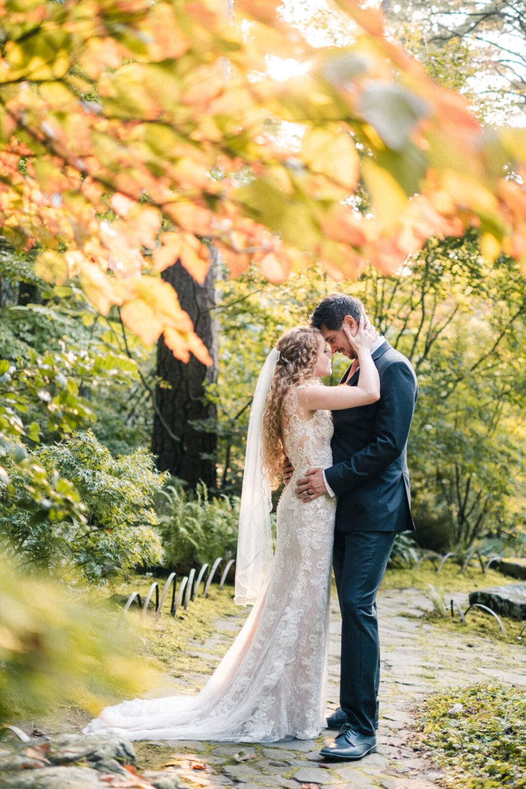 A bride and groom embrace lovingly on a stone path surrounded by lush greenery and soft, warm sunlight filtering through the trees. The bride wears a lace wedding gown and veil, while the groom is dressed in a dark suit.