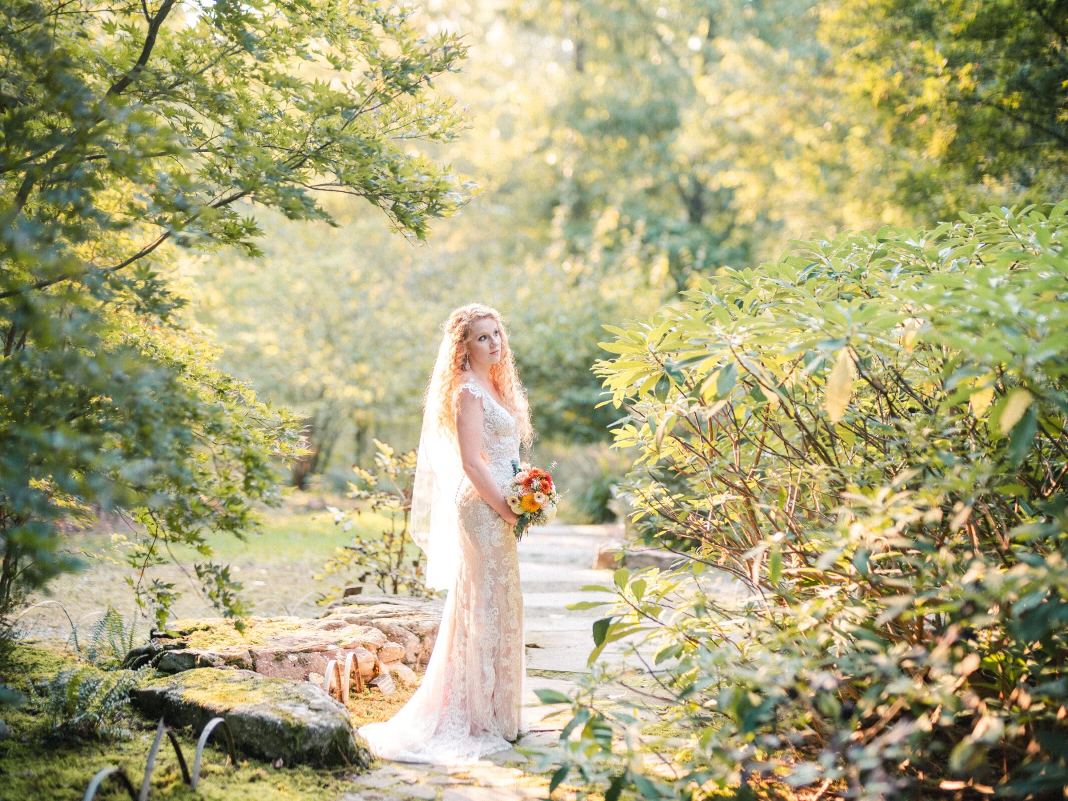 A bride in a lace gown and veil stands on a stone path surrounded by lush greenery, holding a bouquet of orange flowers. Sunlight filters through the trees, creating a serene and picturesque scene.