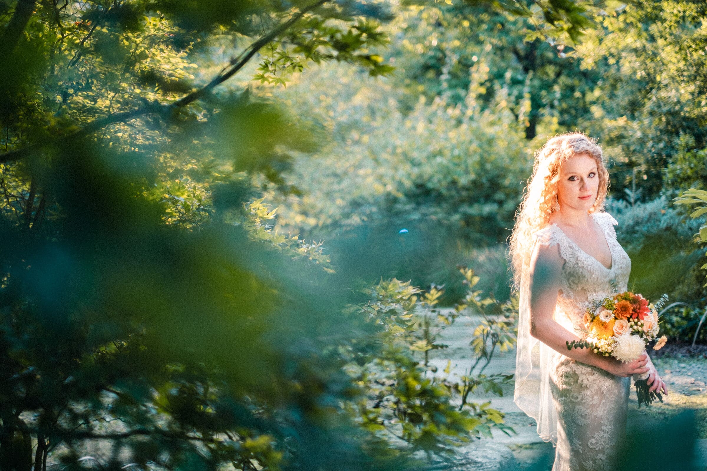 A bride in a lace wedding dress, holding a bouquet of flowers, stands in a sunlit garden surrounded by lush greenery. Her curly hair falls over her shoulders, and she gazes softly into the distance.