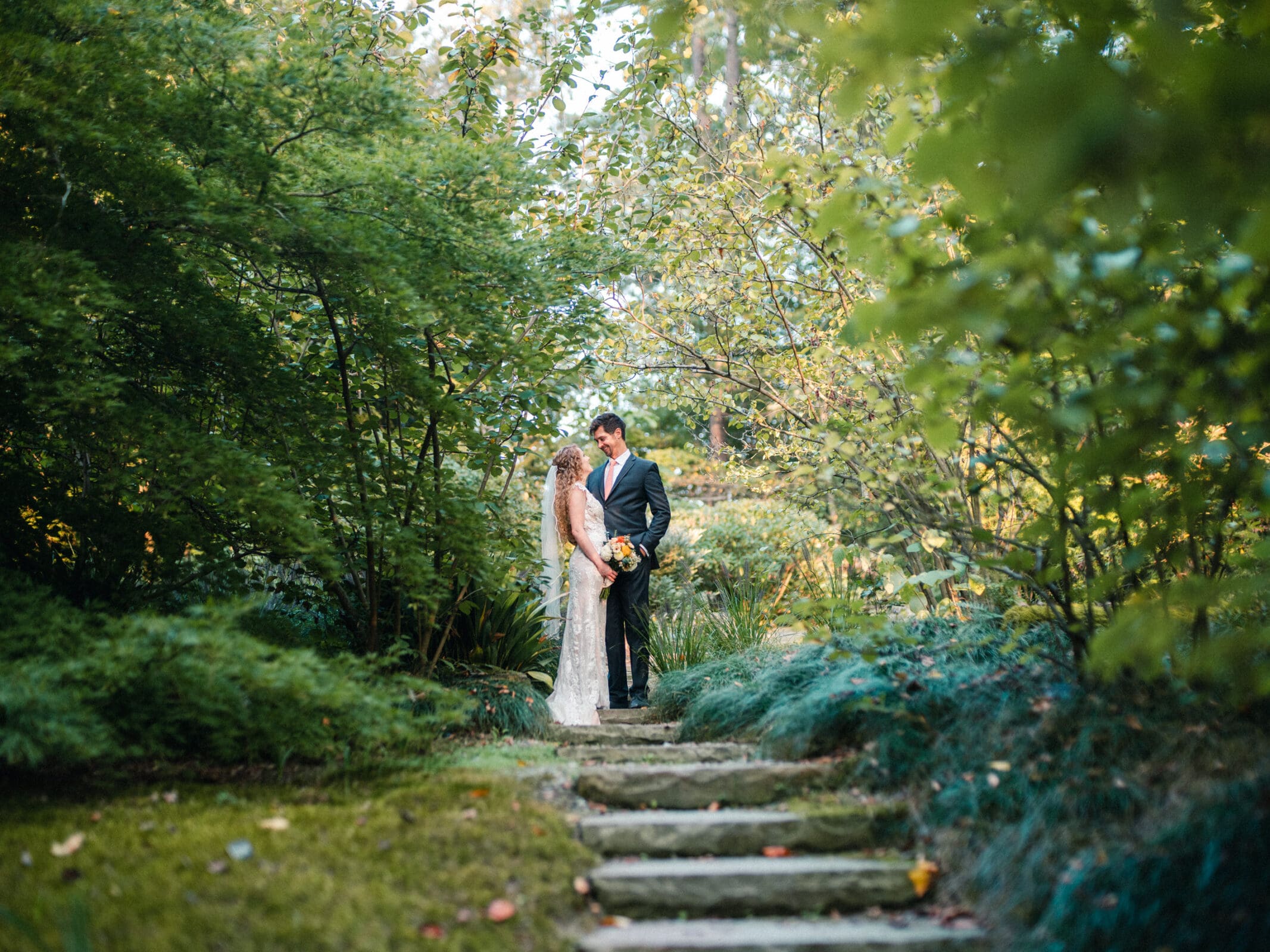 A bride and groom stand closely together on stone steps in a lush garden. The bride holds a bouquet of flowers, and both are surrounded by vibrant green foliage. The light filters softly through the trees.