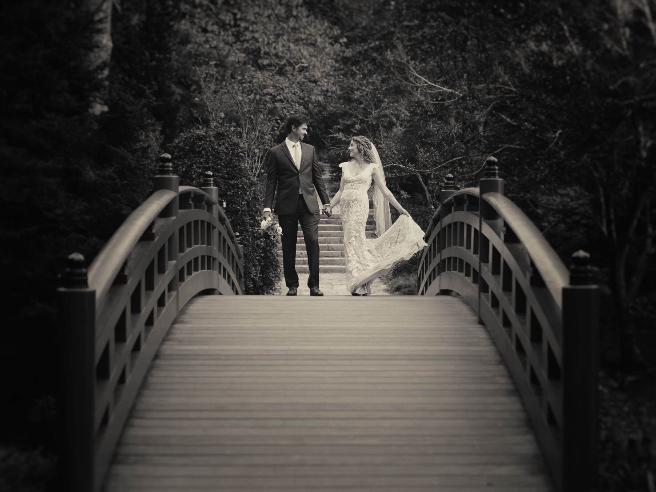 A couple in wedding attire walks hand in hand across a wooden bridge in a forest setting. The scene is captured in black and white, adding a timeless, romantic feel. The bride holds her dress, and the groom carries a bouquet.