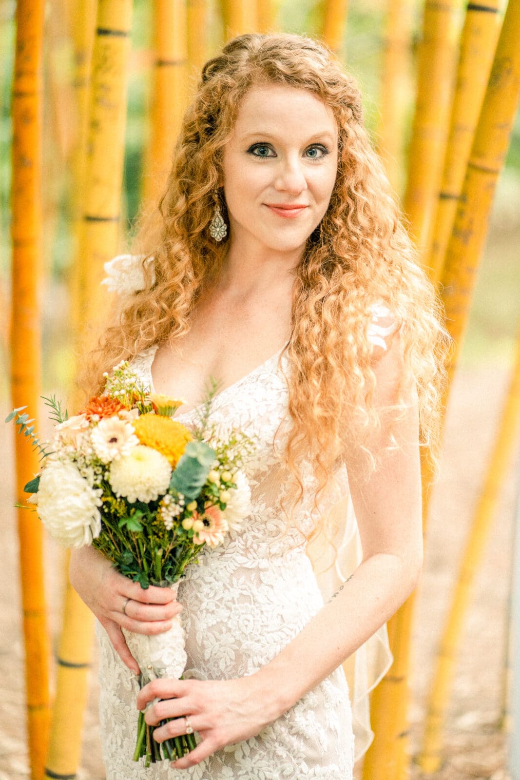 A woman with long curly hair stands in front of bamboo stalks, holding a bouquet of white and yellow flowers. She is wearing a white lace dress and earrings, smiling softly at the camera. The background is lush and green.