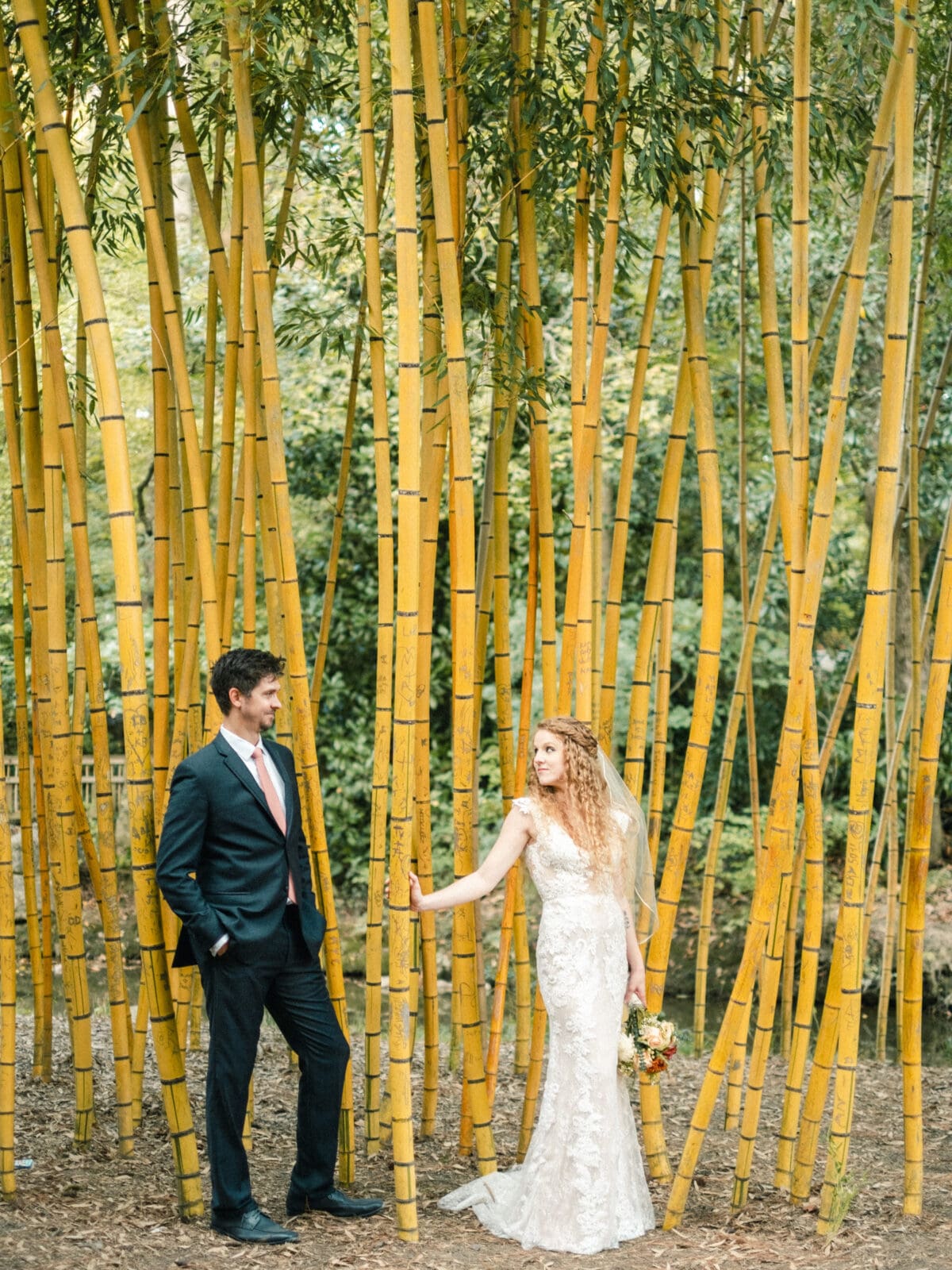 A couple stands among tall yellow bamboo stalks. The woman, in a lace wedding dress, holds a bouquet and looks at the man, who is in a suit with his hands in his pockets. The bamboo grove surrounds them with lush greenery in the background.
